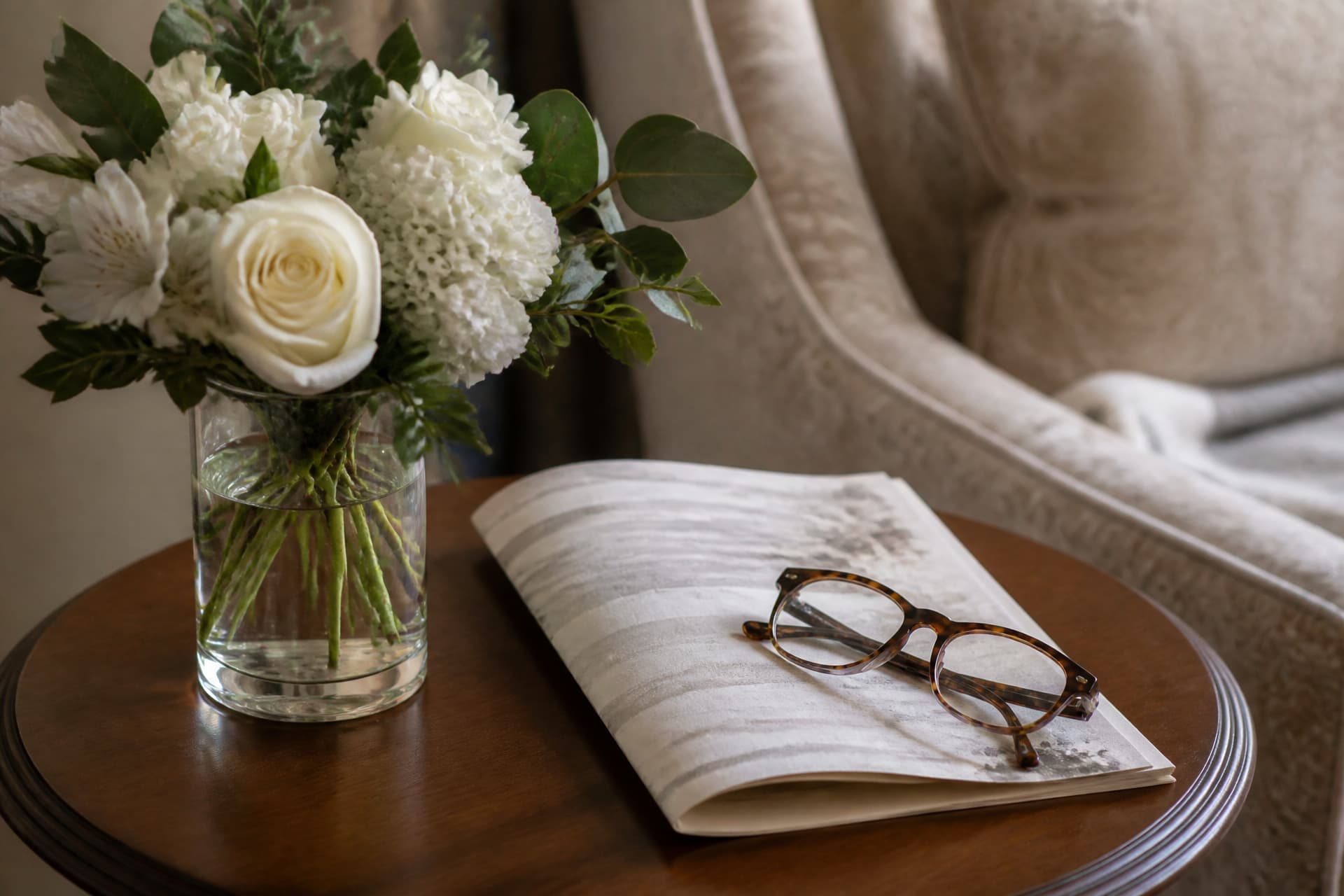 A small arrangement of white flowers on a side table beside a folded printed program and a pair of reading glasses, soft light