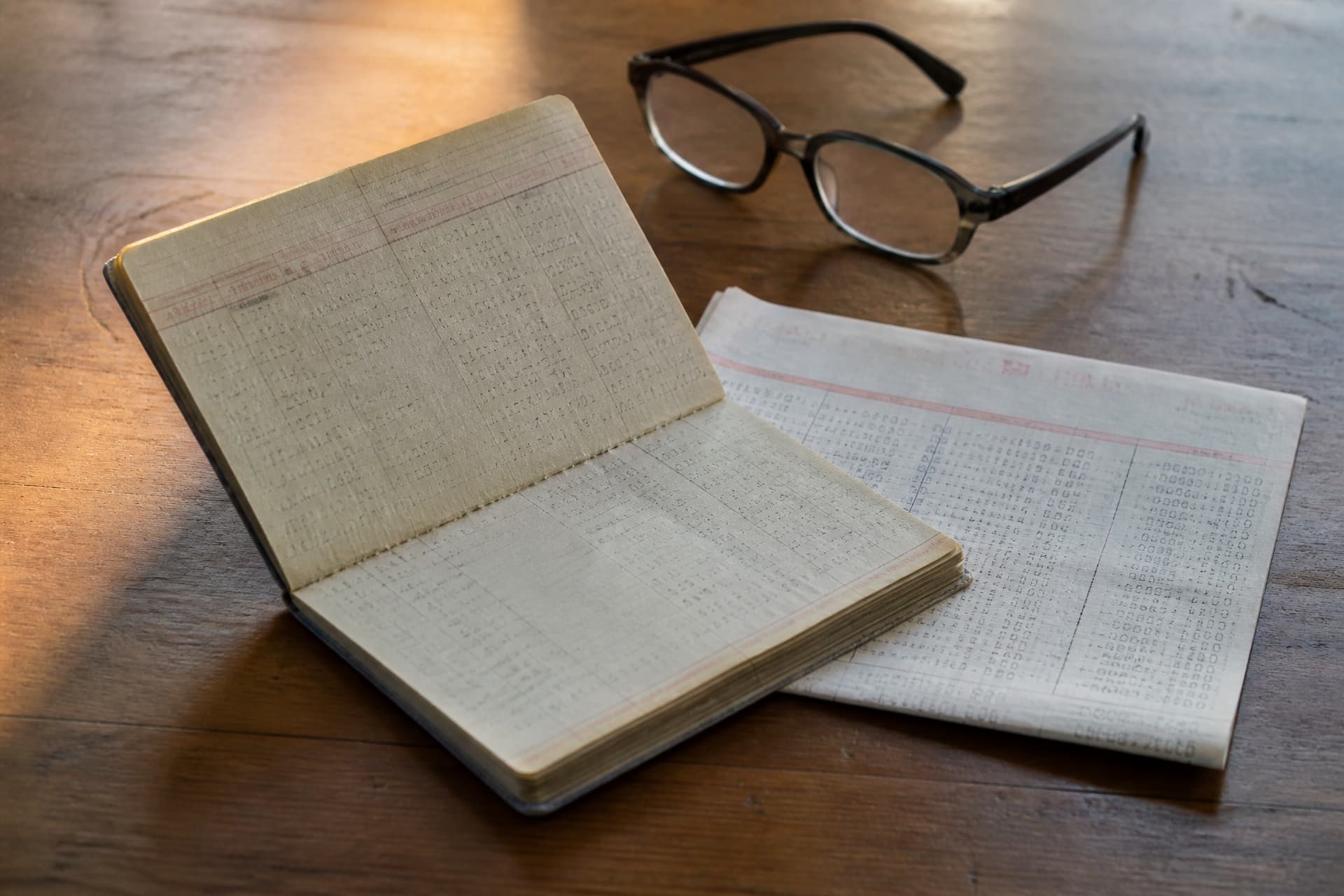 An old savings passbook lying open on a wooden table beside a folded statement and a pair of reading glasses, warm daylight