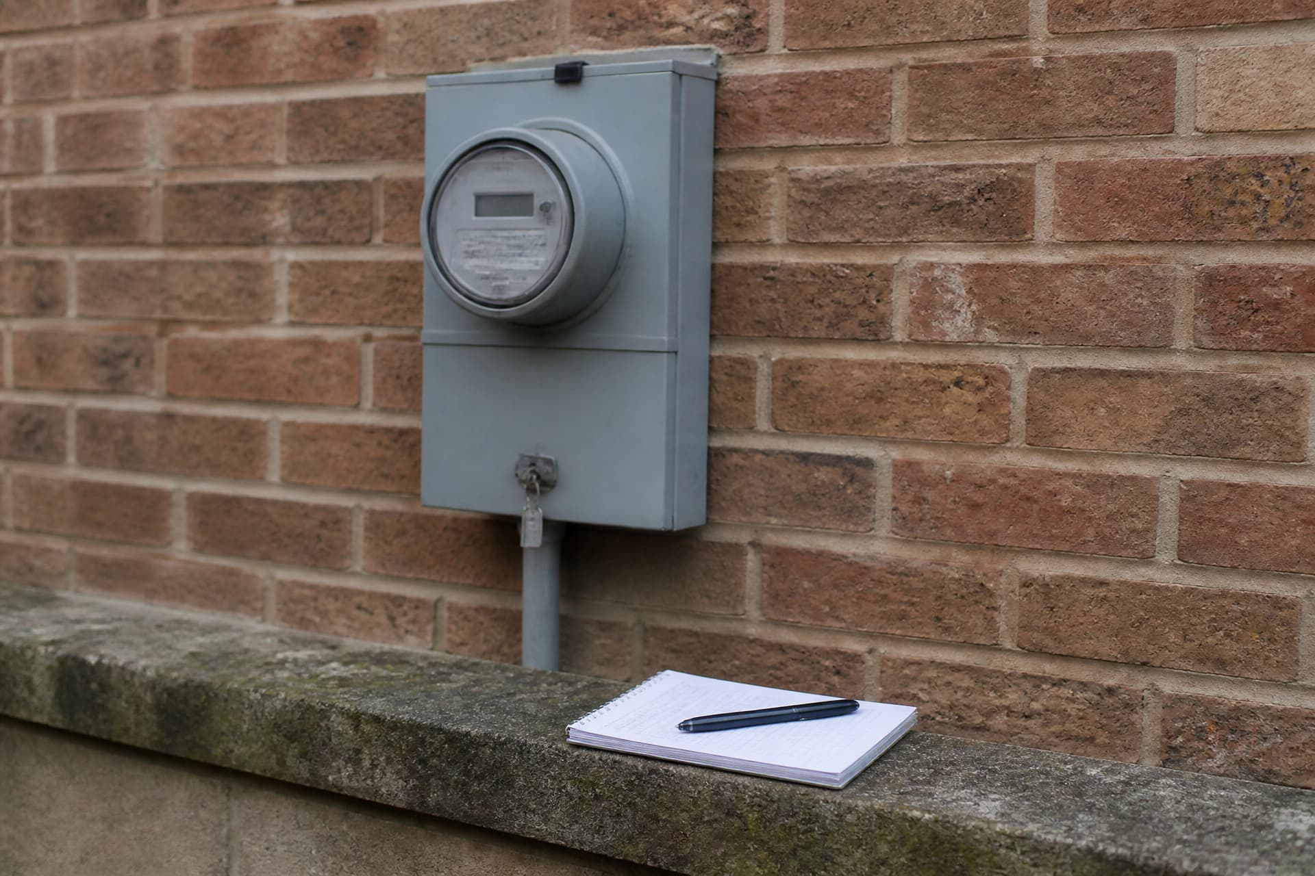 A domestic utility meter mounted on a plain exterior wall with a notebook and pen resting on the ledge below it