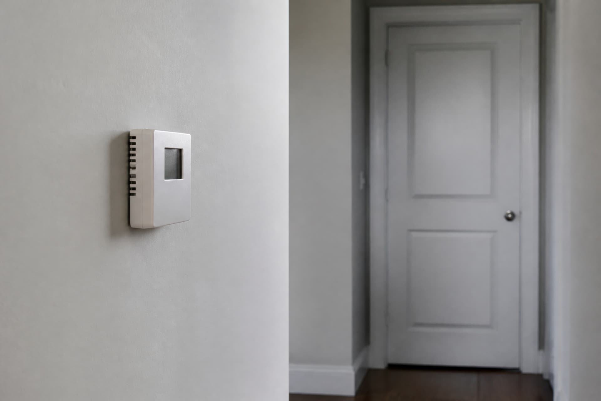 A wall thermostat in an otherwise bare hallway with a closed door beyond it, in cool natural light