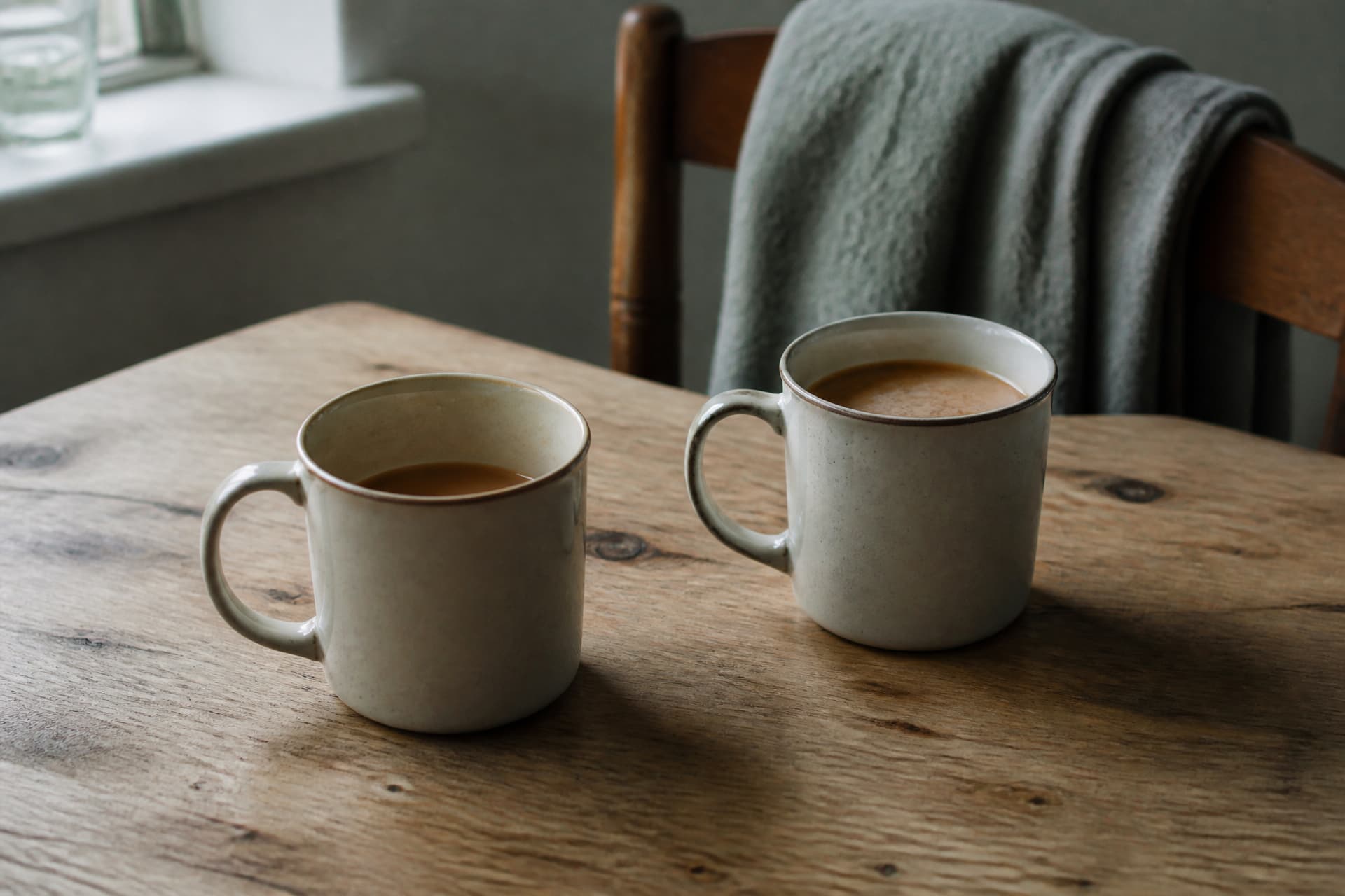 Two ceramic mugs of tea on a scrubbed wooden kitchen table, one half drunk and one untouched, a grey blanket over the chair behind them