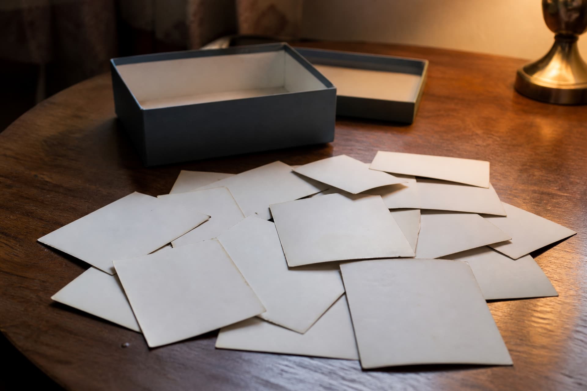 A scatter of old photographs lying face down with only their blank backs showing, beside an open shoebox on a wooden table in lamplight