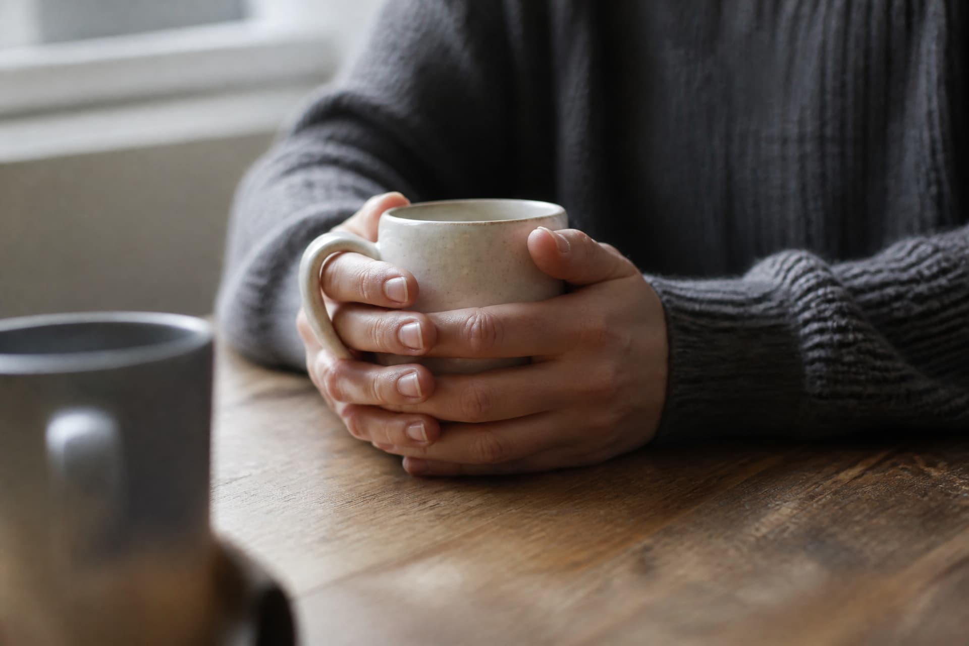 Two hands wrapped around a stoneware mug on a wooden kitchen table with a second mug just in frame opposite, grey daylight from a window