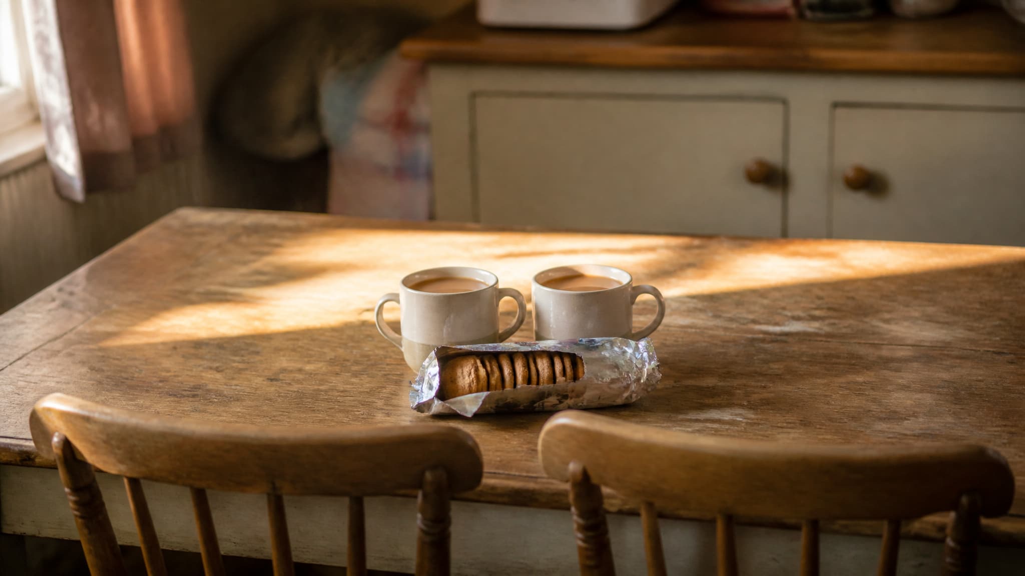 Two mugs on a kitchen table beside an unopened packet of biscuits in soft afternoon light, two chairs pulled close together