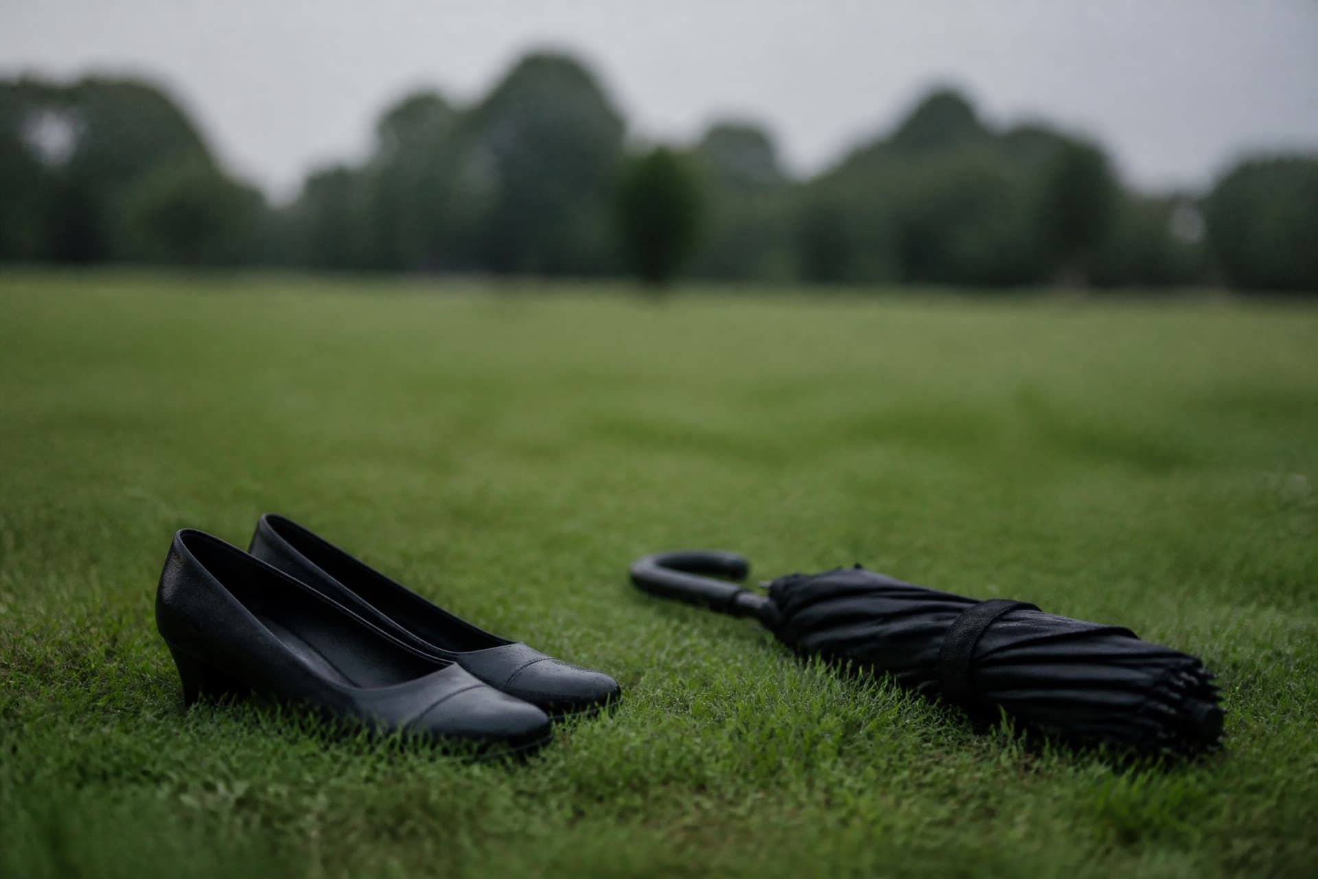 Plain black low-heeled shoes standing on wet grass beside a folded dark umbrella at a graveside service on an overcast day