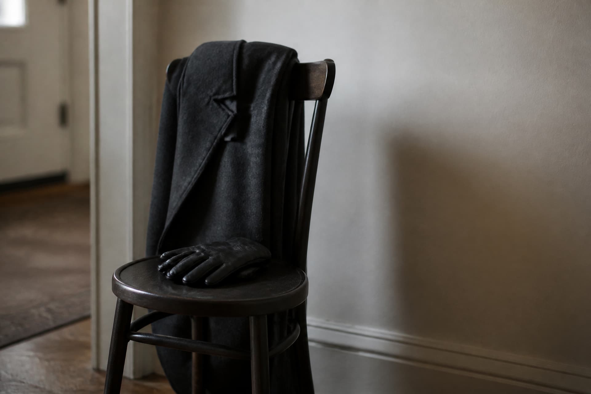 A dark wool overcoat folded over the back of a hall chair with a leather glove resting on the seat
