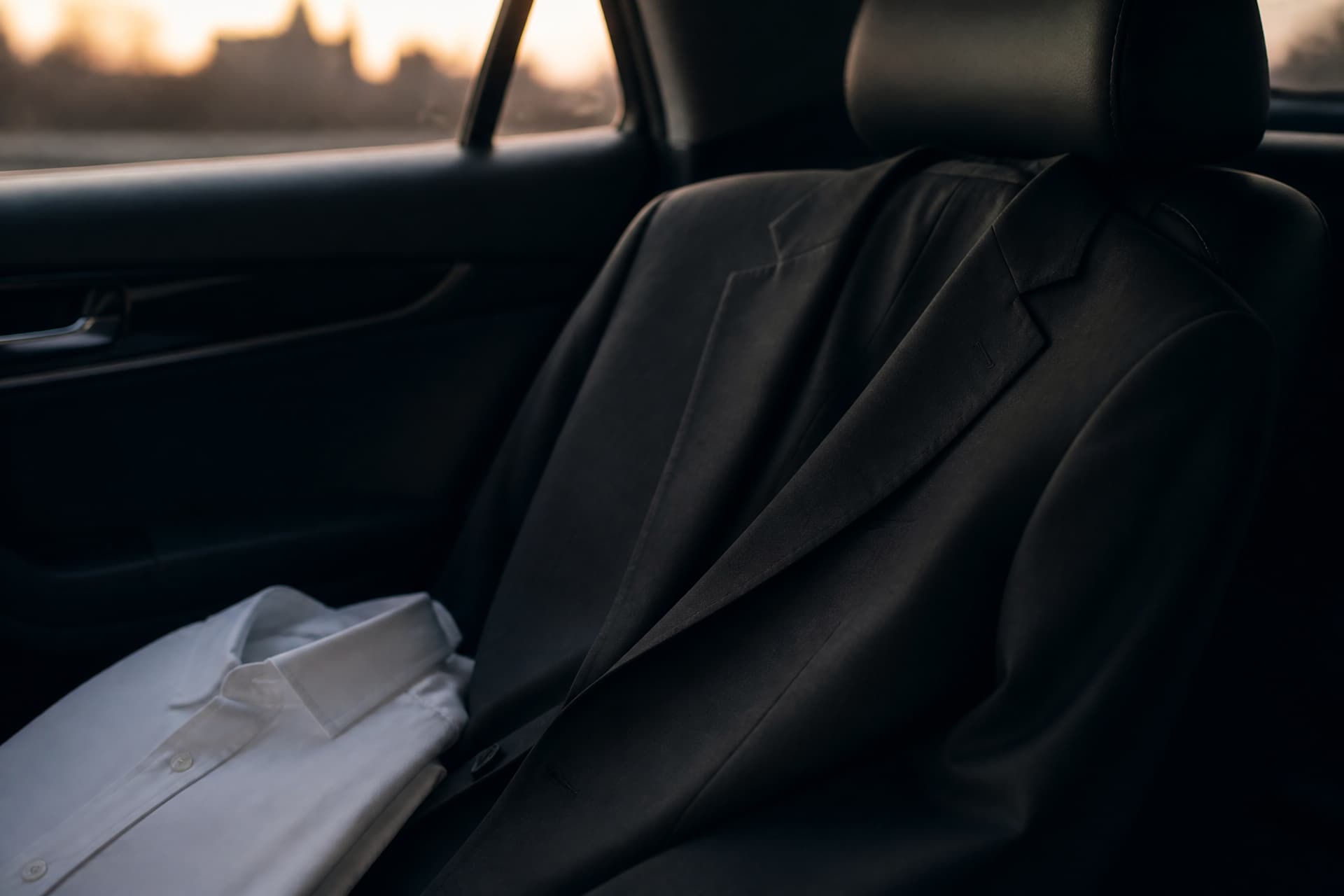 A dark jacket folded over the back of a car seat beside a plain collared shirt, in low evening light