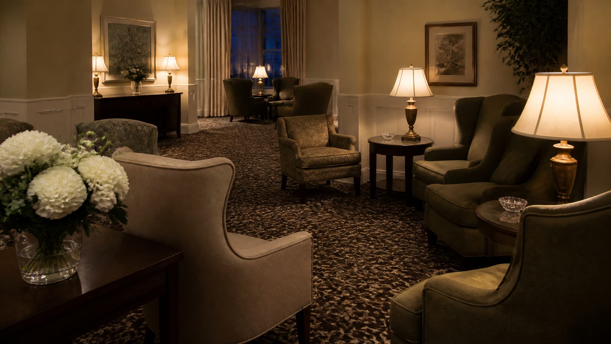 A quiet funeral home reception room with lamps lit and empty upholstered chairs arranged in small groups