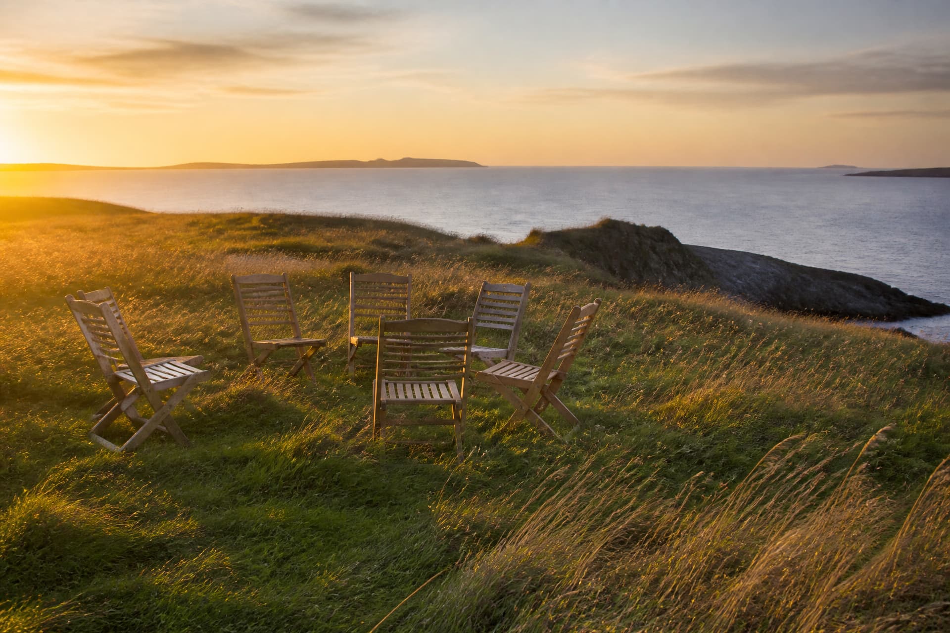Wooden folding chairs arranged in a loose circle on a grassy headland above the sea at golden hour
