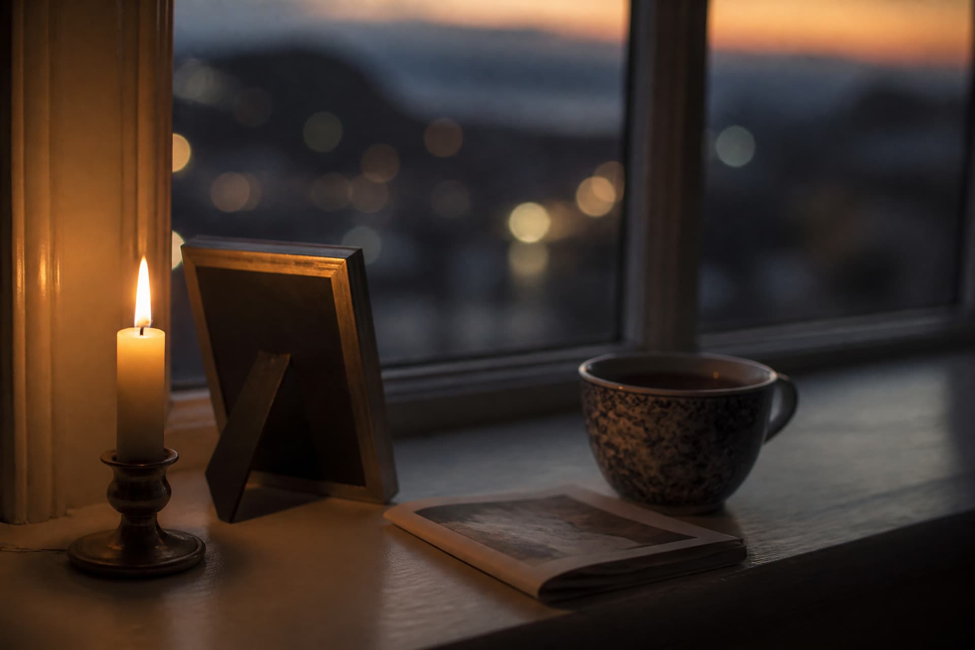 A single lit candle on a windowsill beside a small framed photograph turned away from the camera, a mug and a folded program on the sill