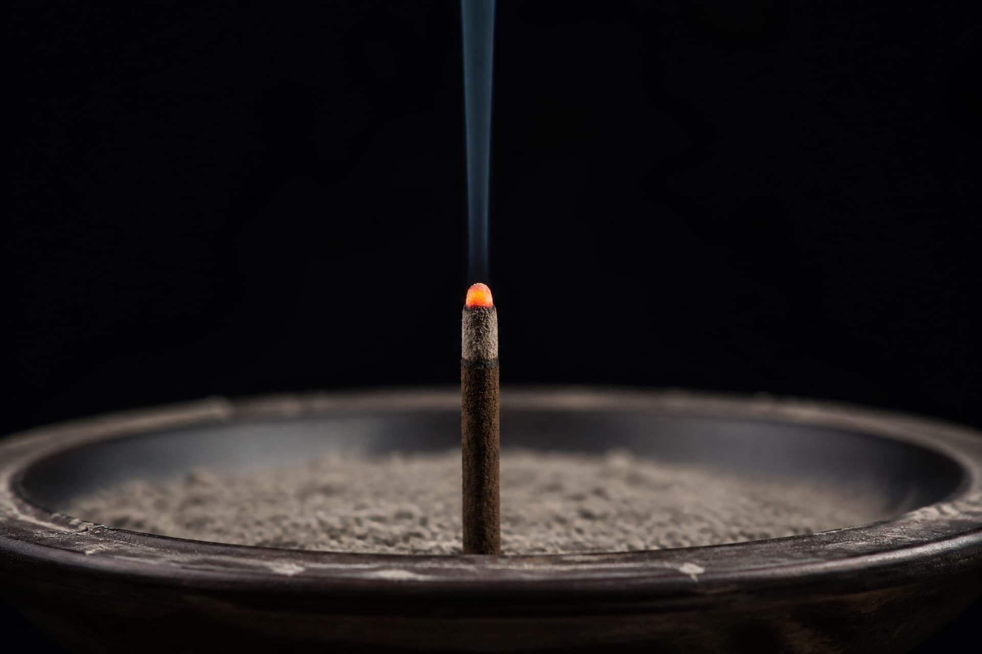 A single stick of incense burning down in a bowl of gray ash, very close, dark background, one thin line of smoke rising