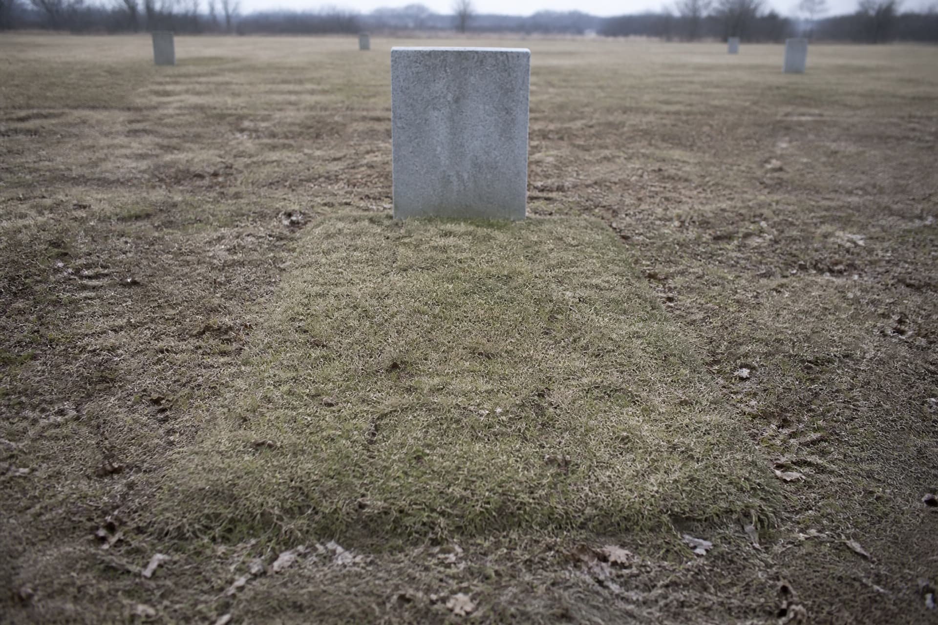 A simple raised earth grave in a quiet cemetery with a plain unlettered upright stone at its head, dry grass around it under overcast light