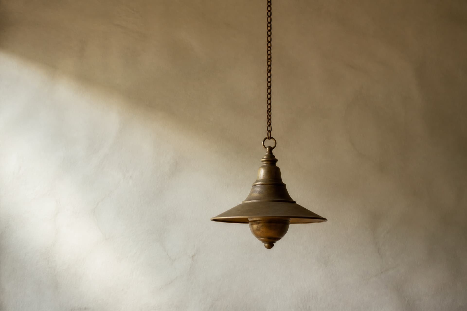 A single unlit hanging brass lamp on a long chain against a plain lime-washed wall, lit softly by daylight from one side