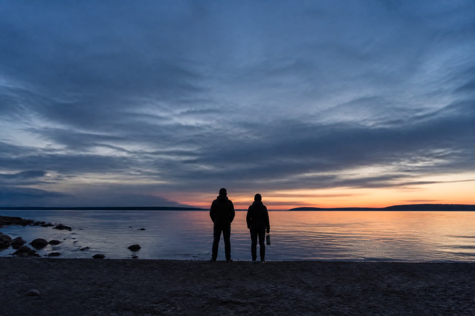 Two people standing at a shoreline at dusk, seen from behind at a distance, one holding a small cylindrical container