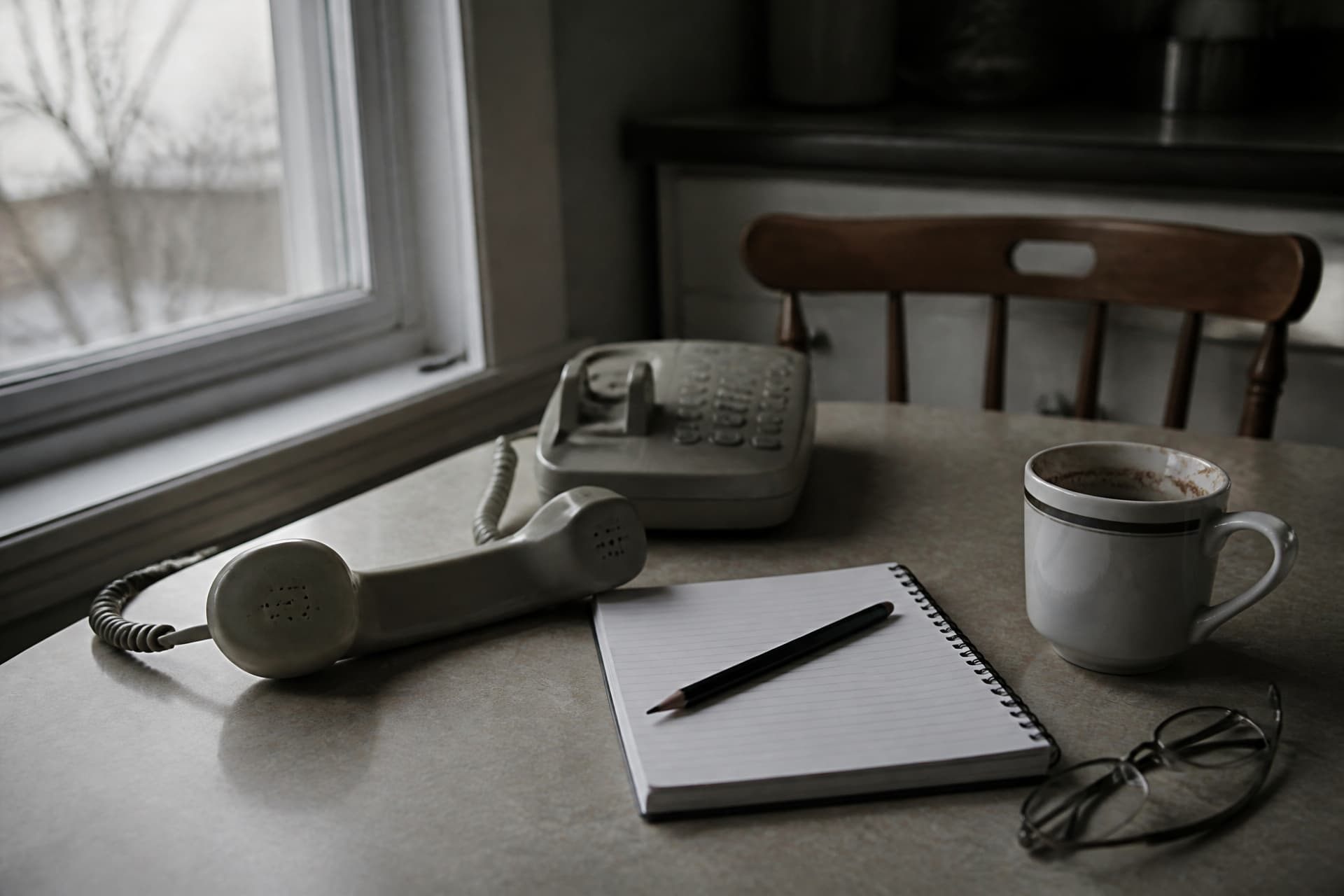 A kitchen table by a window on an overcast morning with a landline handset off its cradle, a spiral notepad open at a blank page with a pencil across it, a half-finished mug of coffee and folded reading glasses