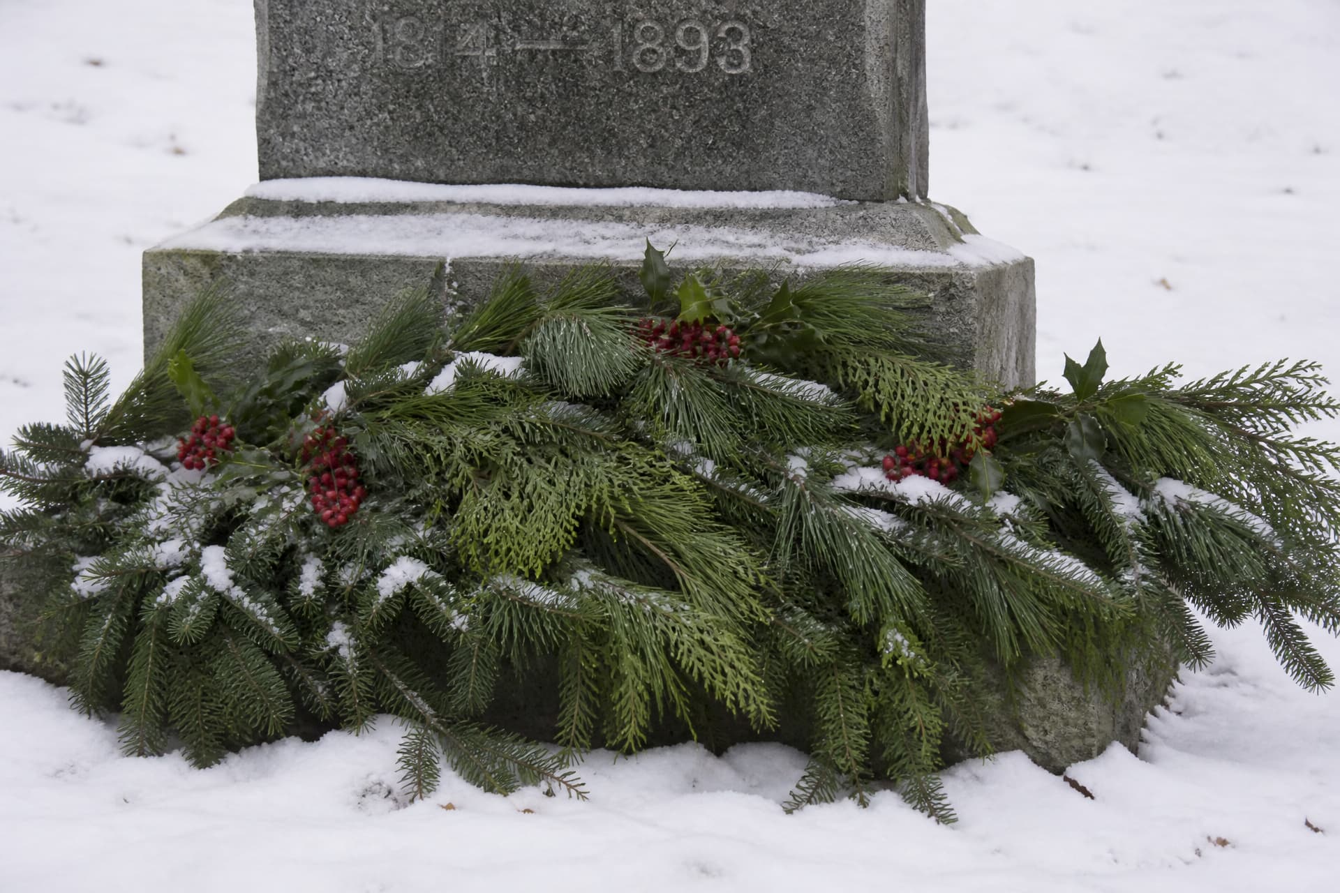 A granite headstone base in winter with a spray of cut pine, cedar and fir boughs laid across it alongside sprigs of holly heavy with red berries, fresh snow on the ground and dusting the needles