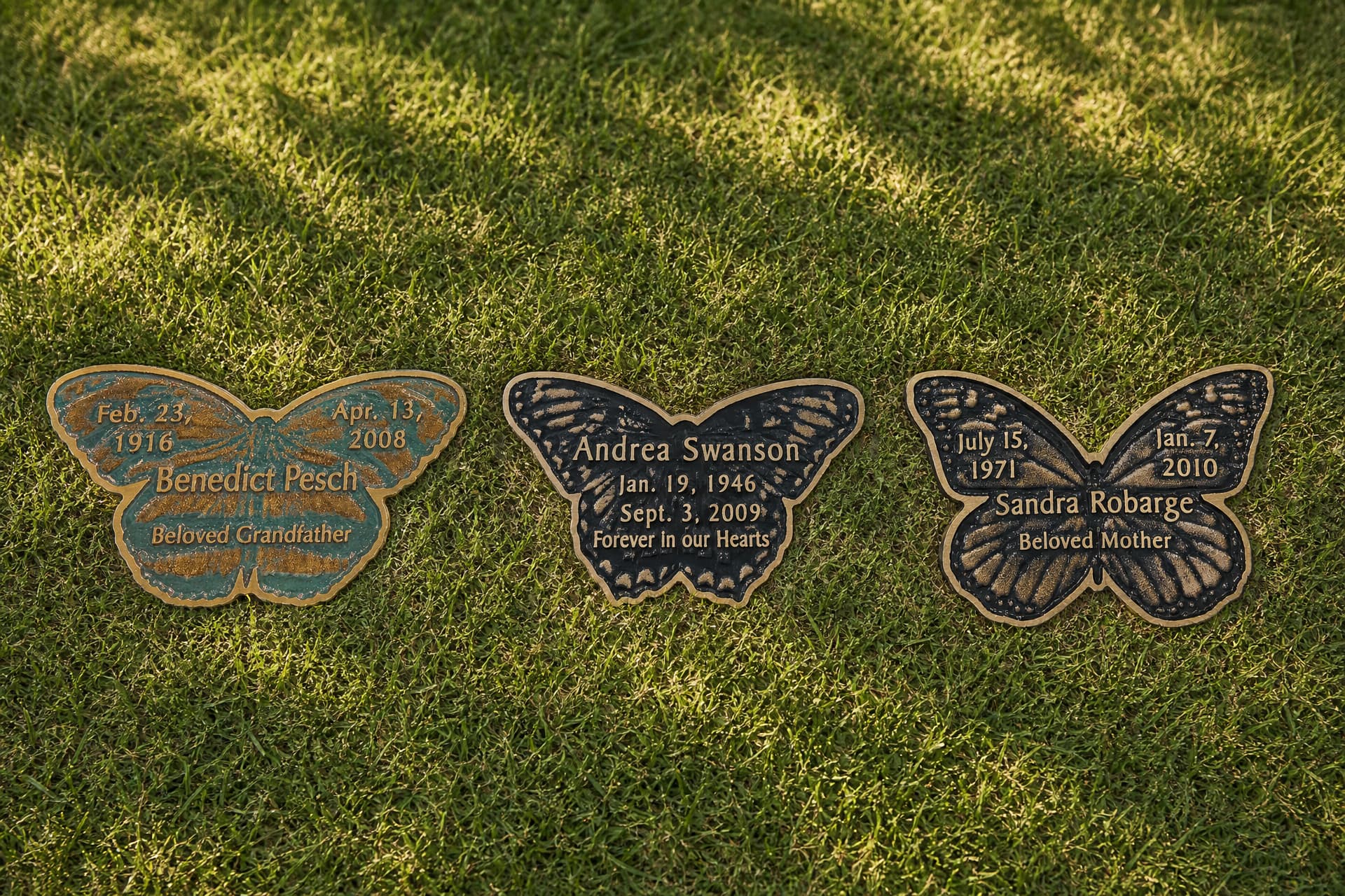 Three butterfly-shaped cast bronze grave markers in green, black and bronze finishes set flush in mown cemetery grass, each carrying a name and dates