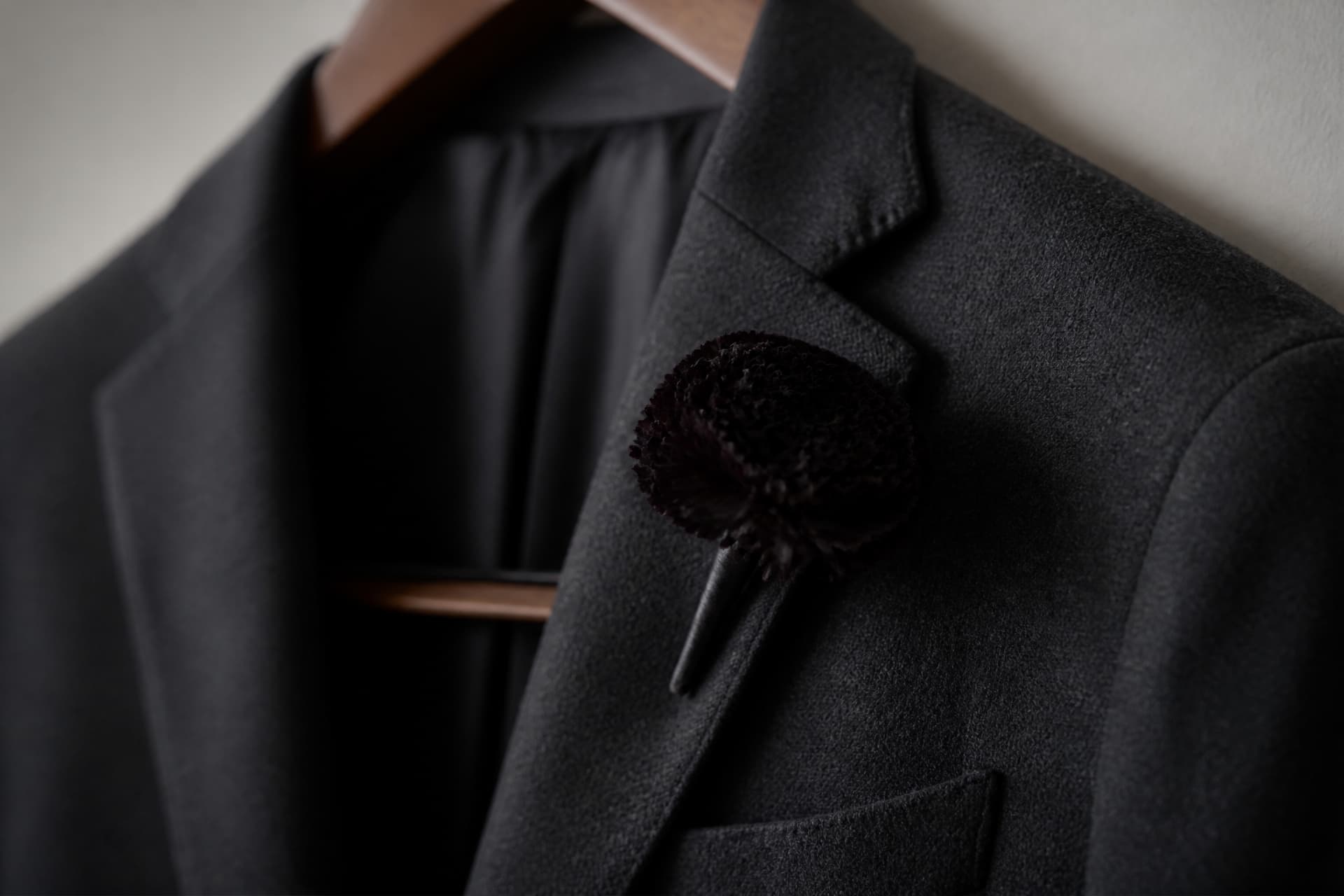 A single dark carnation boutonniere pinned to the lapel of a charcoal wool suit jacket hanging on a wooden hanger against a plain wall