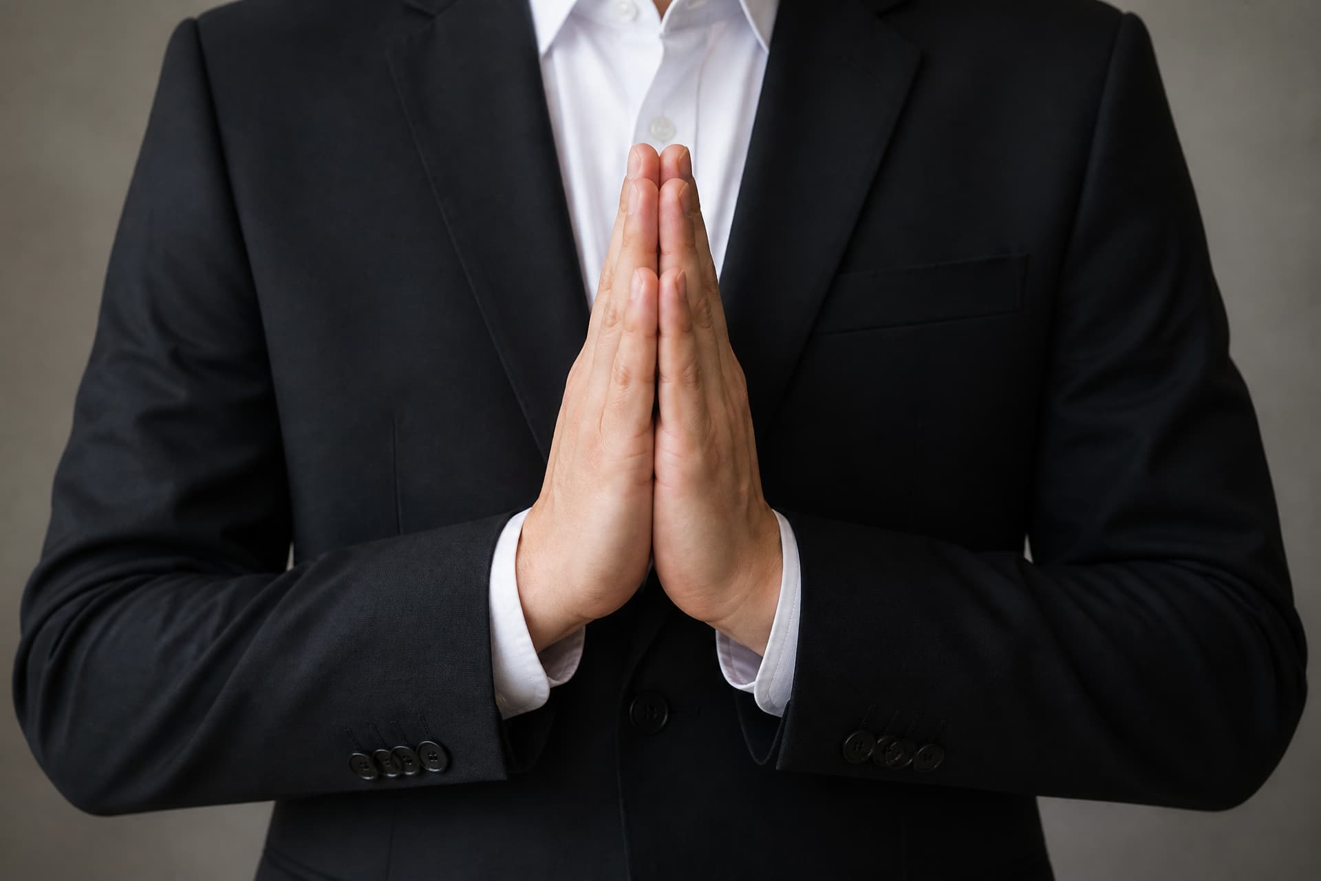 A pair of hands held together in a gesture of respect in front of dark formal clothing