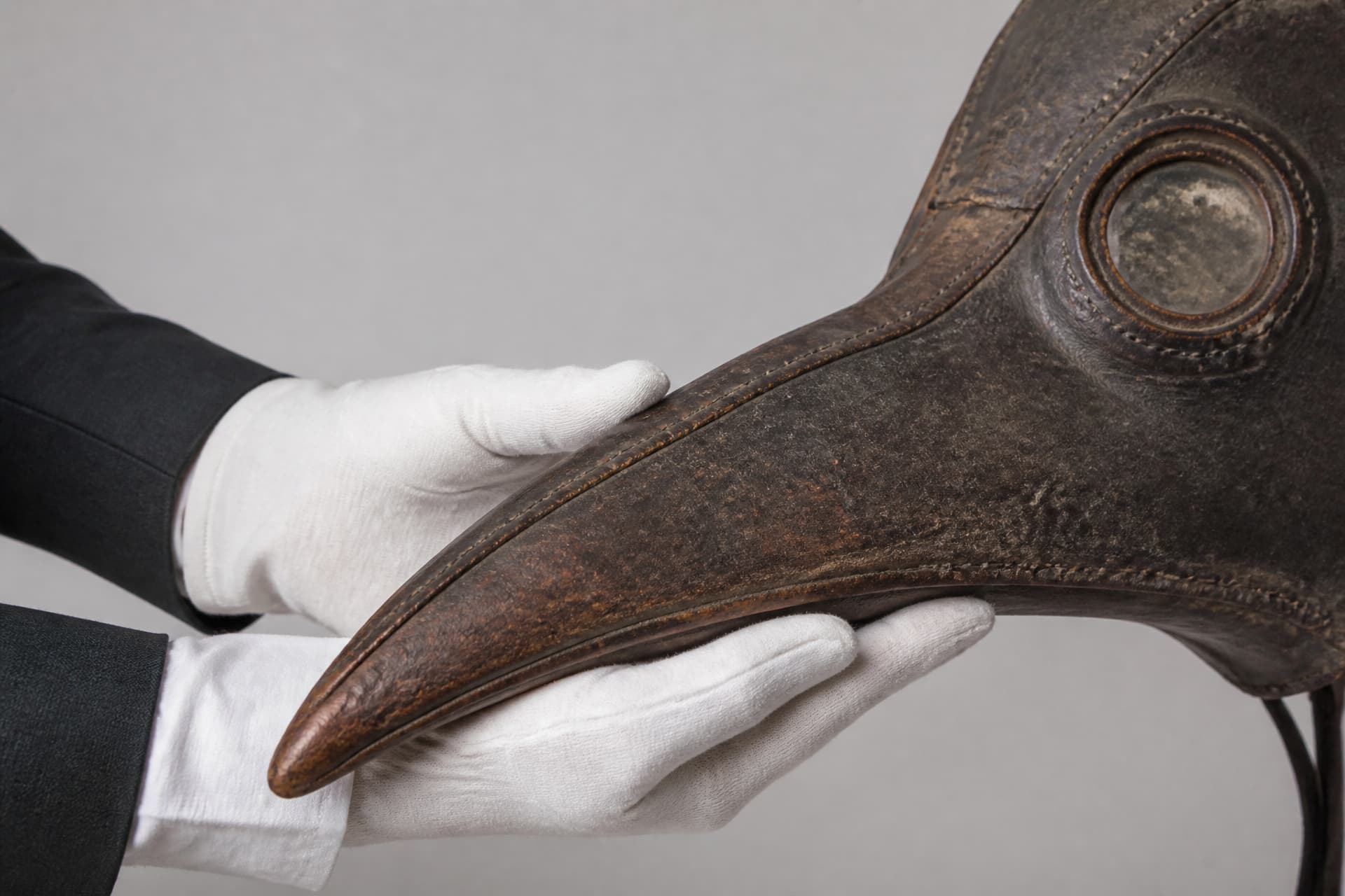 A close-up of a conservator's hands in white cotton gloves supporting the beak of an aged leather beaked mask, with visible stitched seams and a round glass eyepiece