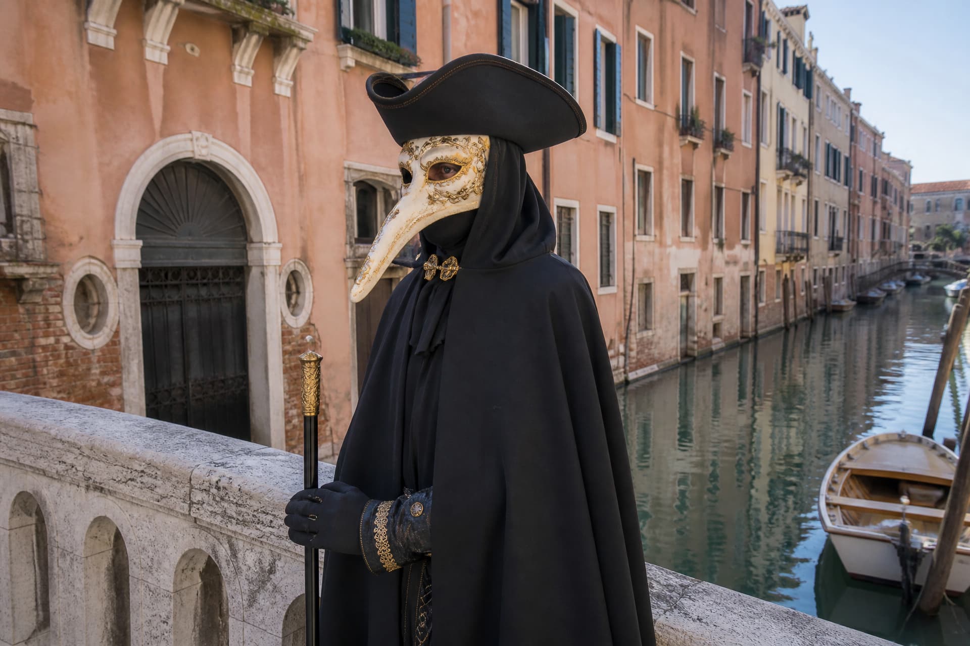 A person in a dark cloak and black tricorn hat wearing a white and gilded Venetian carnival long-beaked mask, standing on a canal bridge in Venice in flat winter daylight with pastel buildings behind