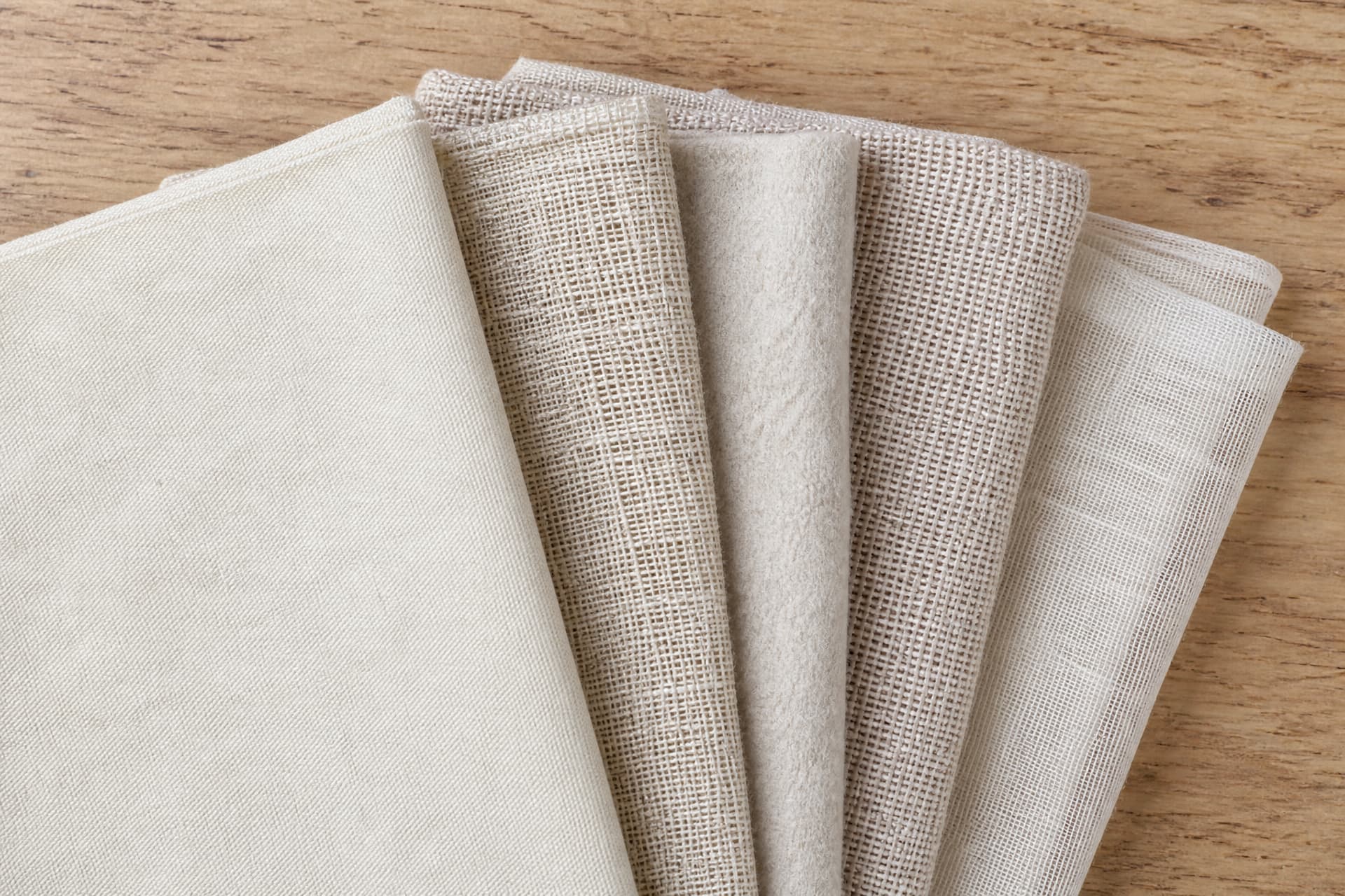 Five folded lengths of undyed natural cloth fanned out on a plain wooden surface, each one a different weave, seen from directly above