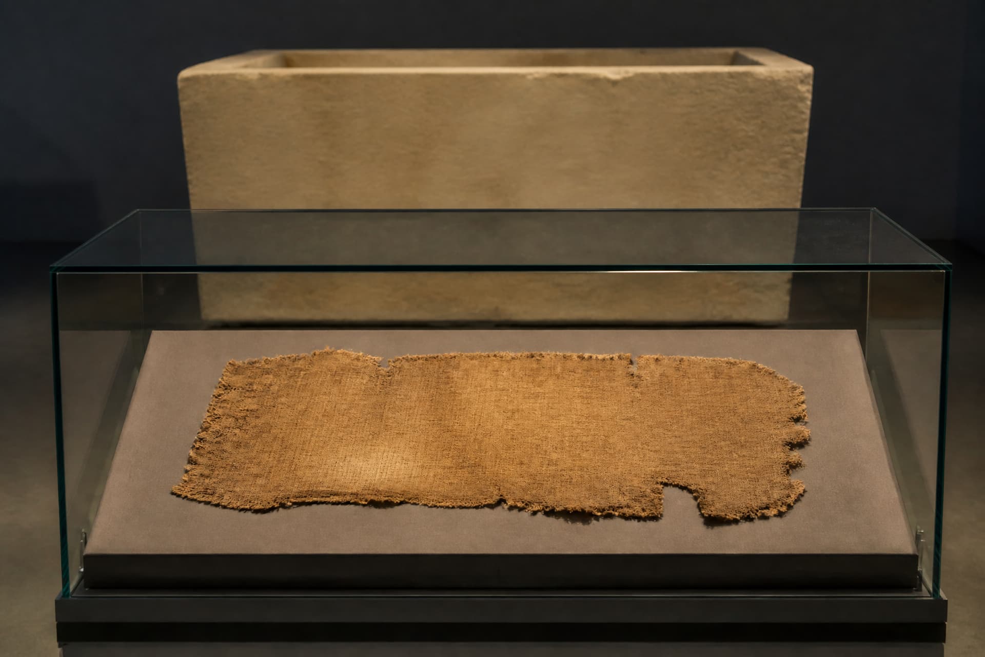A museum vitrine holding a fragment of ancient undyed linen wrapping on a neutral gray mount, with the blurred edge of a stone sarcophagus behind it