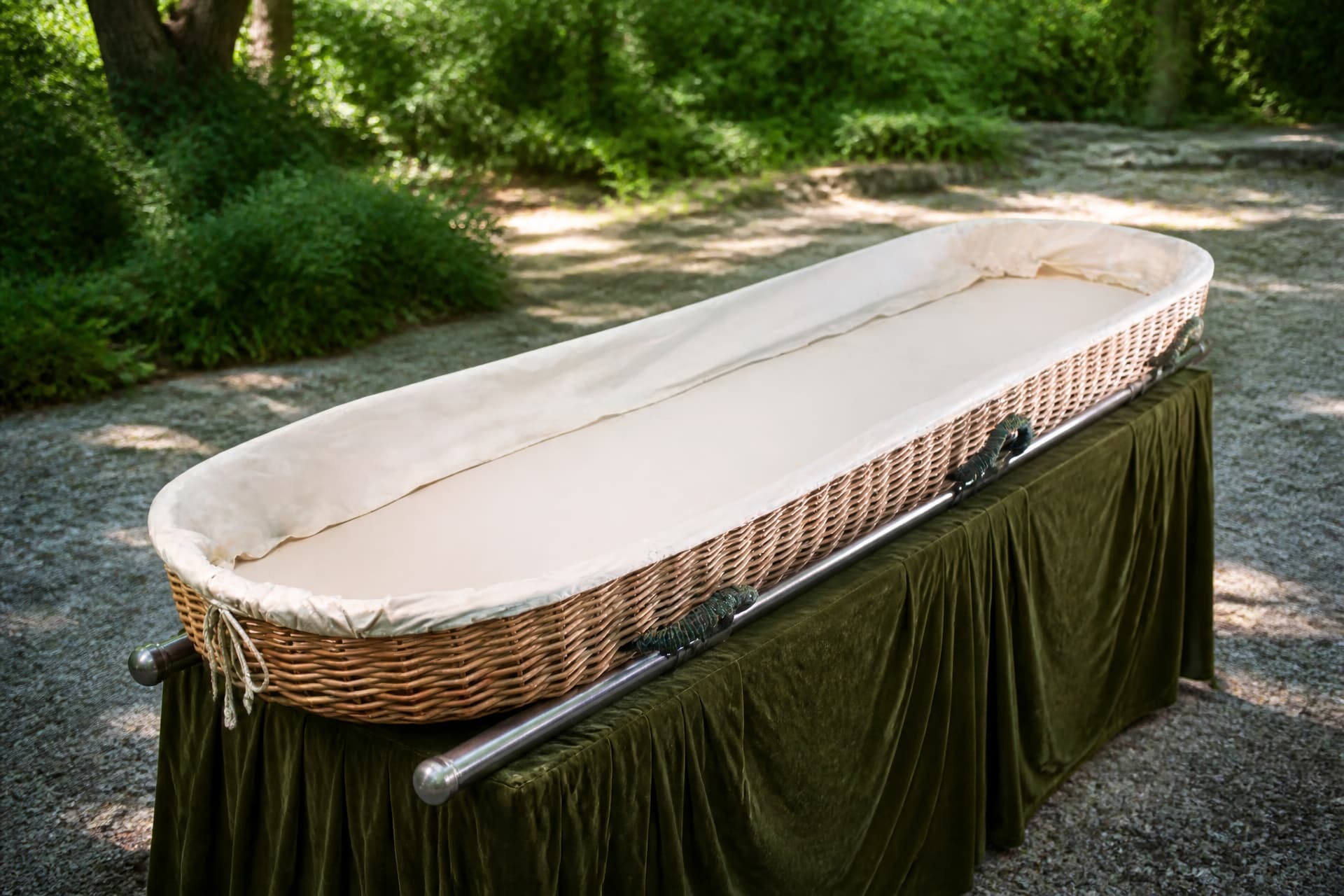 A shallow oval woven willow carrying tray lined with cream cotton, empty, resting on a draped bier on a gravel path with greenery behind it