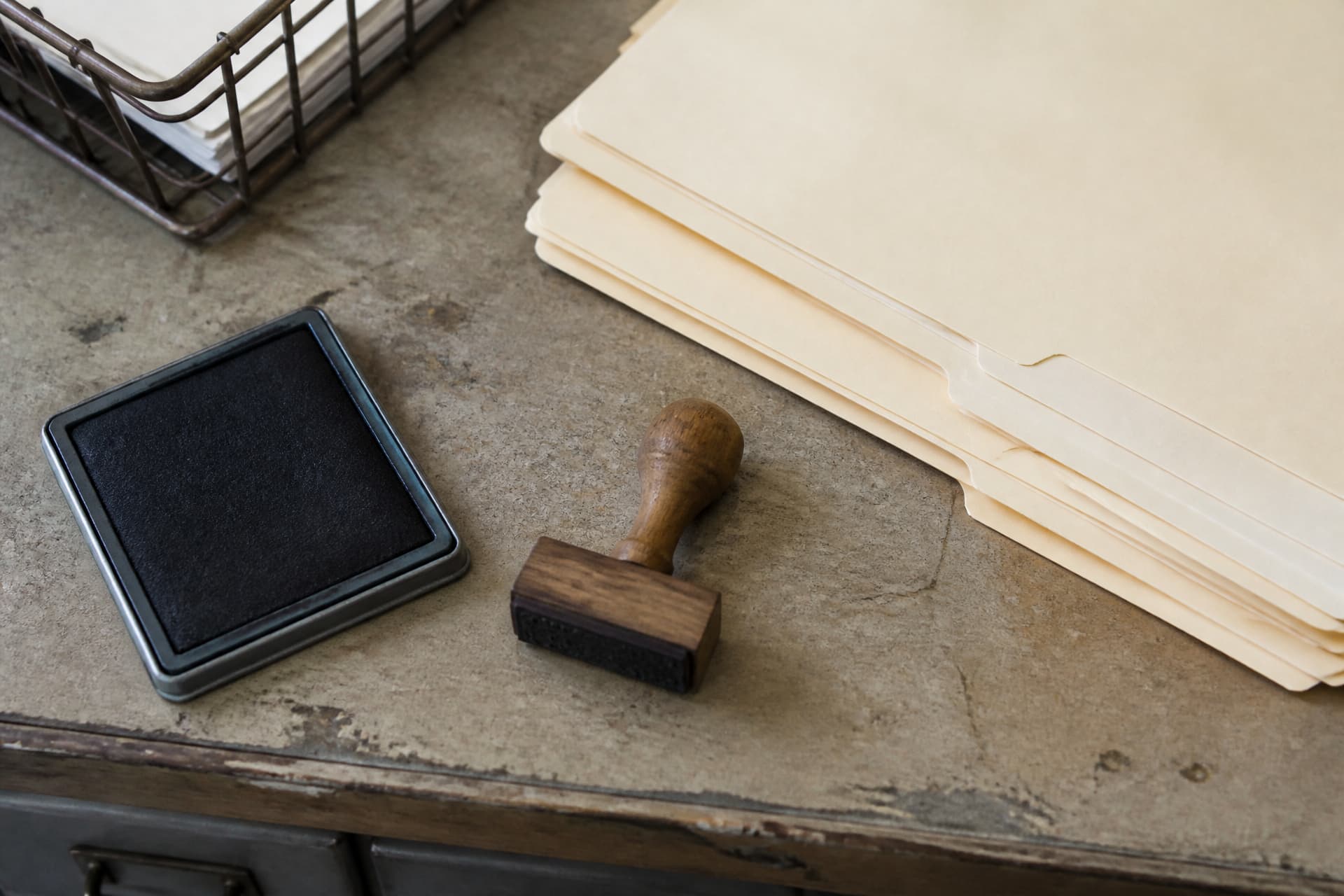 A close view of a rubber date stamp and ink pad beside a stack of pale manila folders on a worn municipal counter, lit by flat window daylight, with a wire filing tray at the edge of the frame