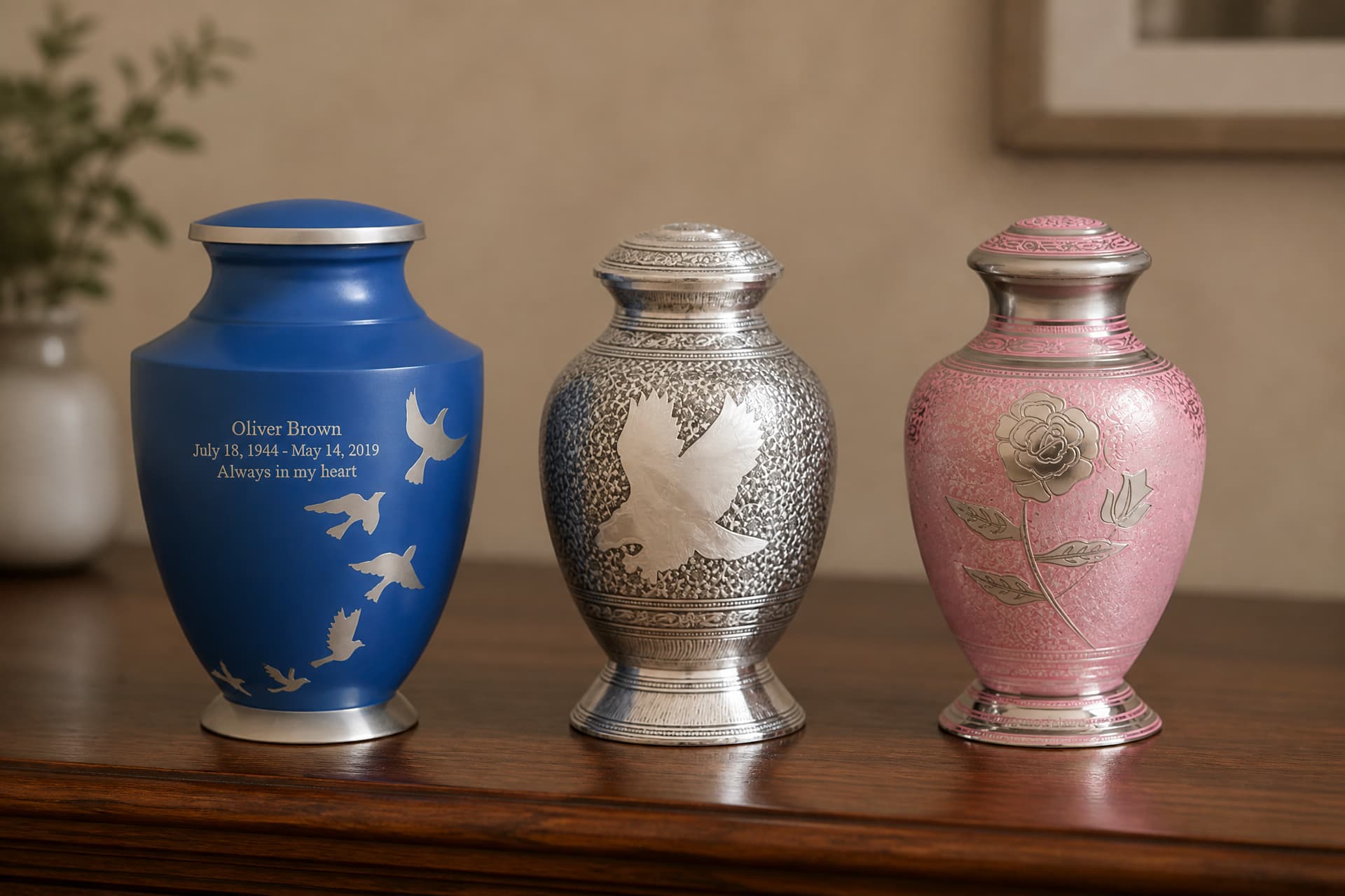 Three full-size cremation urns of different materials and profiles arranged on a plain pale surface in soft even daylight, a brushed metal vase form, a dark wooden box and a lighter stone-finish urn