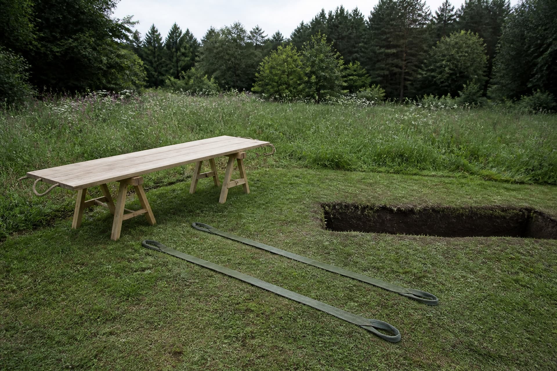 A plain wooden bier board with rope handles resting on trestles beside an open grave in a meadow burial ground, two empty webbing lowering straps laid out flat on the grass alongside