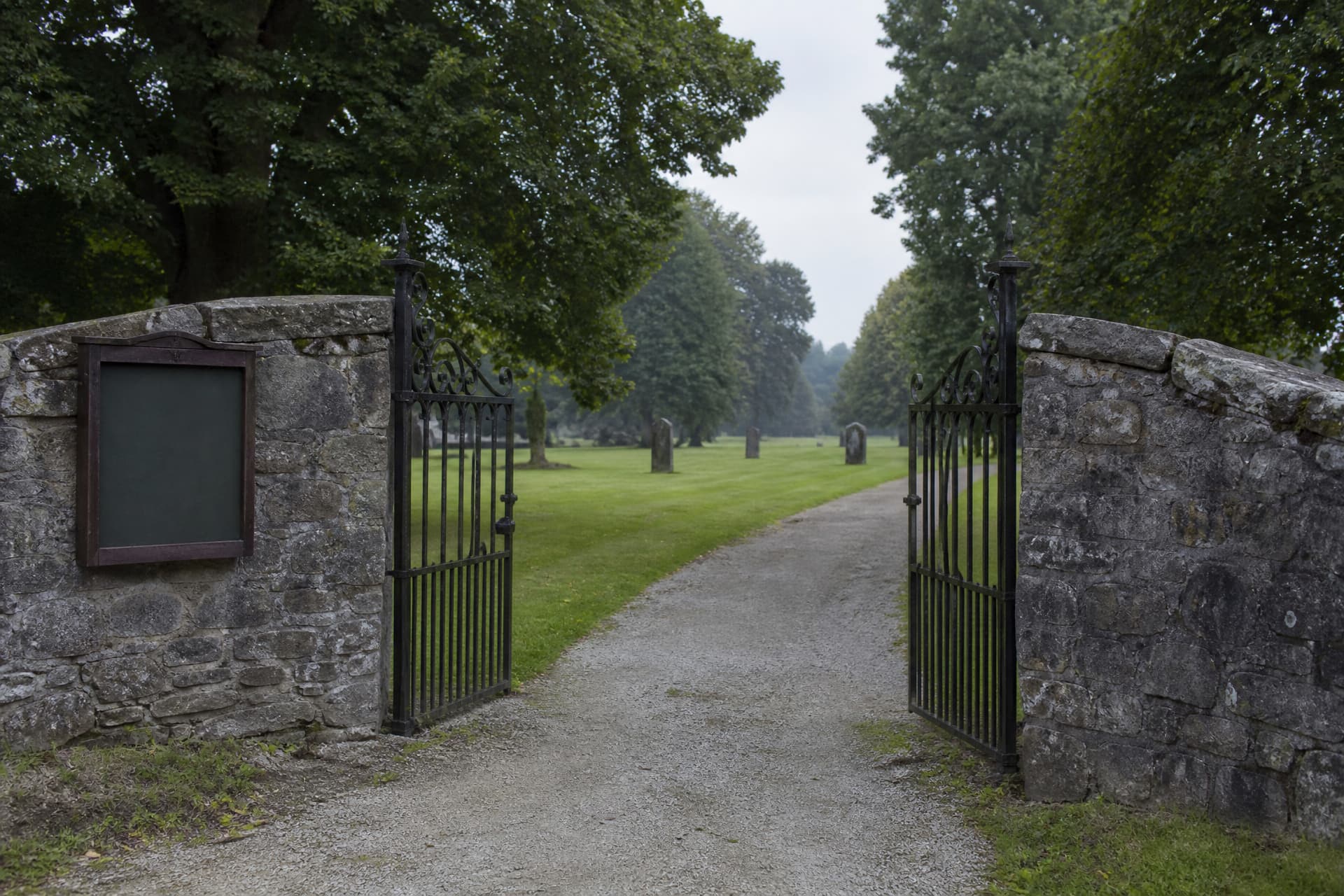 A wrought iron cemetery gate standing open on a gravel path with a low stone wall running away on both sides, mown grass and mature trees beyond, and an empty noticeboard frame mounted on the wall