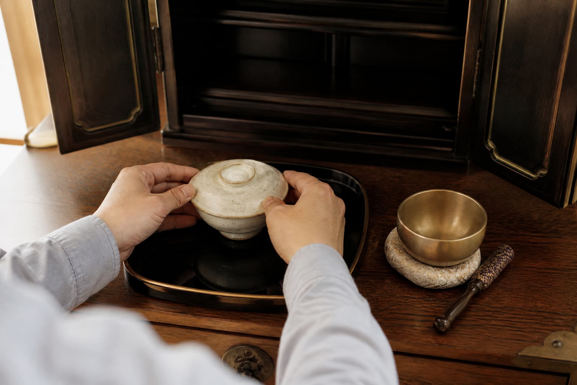 Close over-the-shoulder view of two hands setting a small covered rice bowl onto a low tray in front of an open lacquered cabinet shrine, with a brass bowl-bell and striker beside it