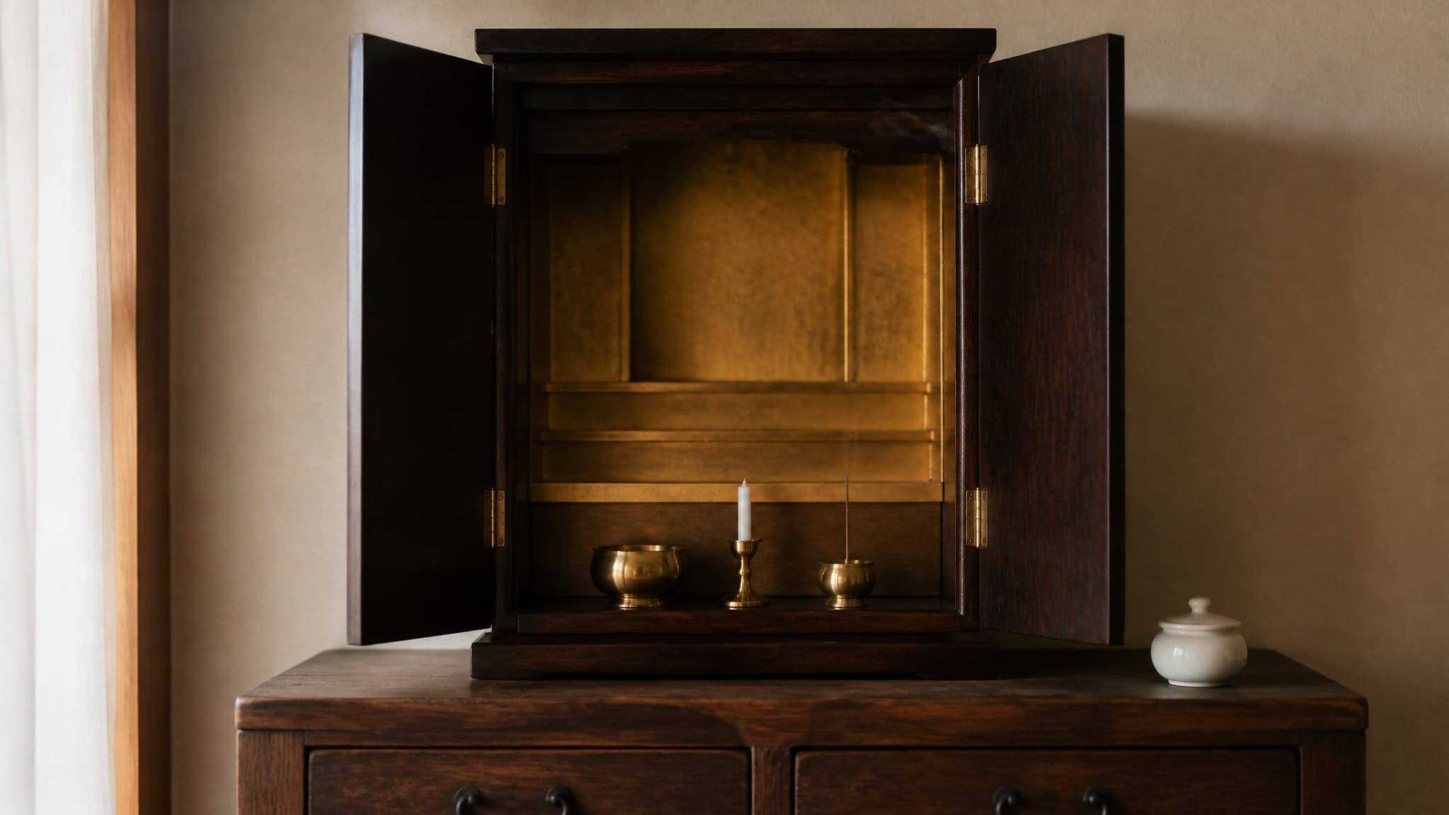 A dark lacquered wooden cabinet shrine with its doors open against a plain interior wall in soft morning light, holding a low offering bowl, a slim brass candle stand and a single stick of incense