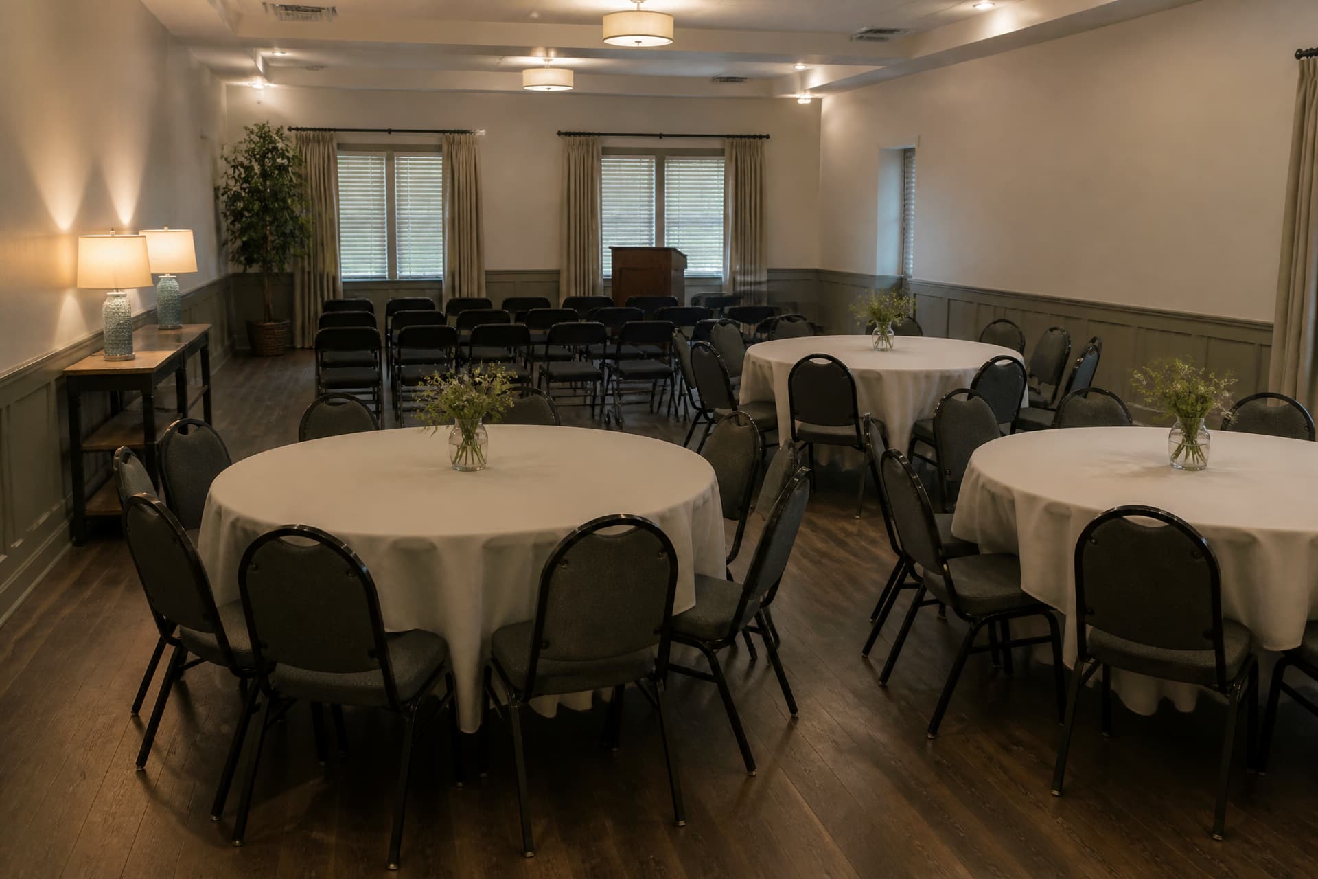 A function room set up before anyone arrives, with three round tables under pale linen cloths and small jars of flowers, a short row of folding chairs facing a low lectern, and two table lamps brought from home on a side table