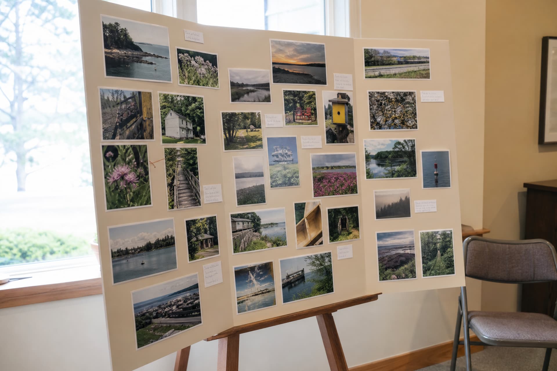 A large tri-fold display board on an easel in a bright room, covered with dozens of photographs in loose clusters and a few small handwritten labels, a folding chair beside it