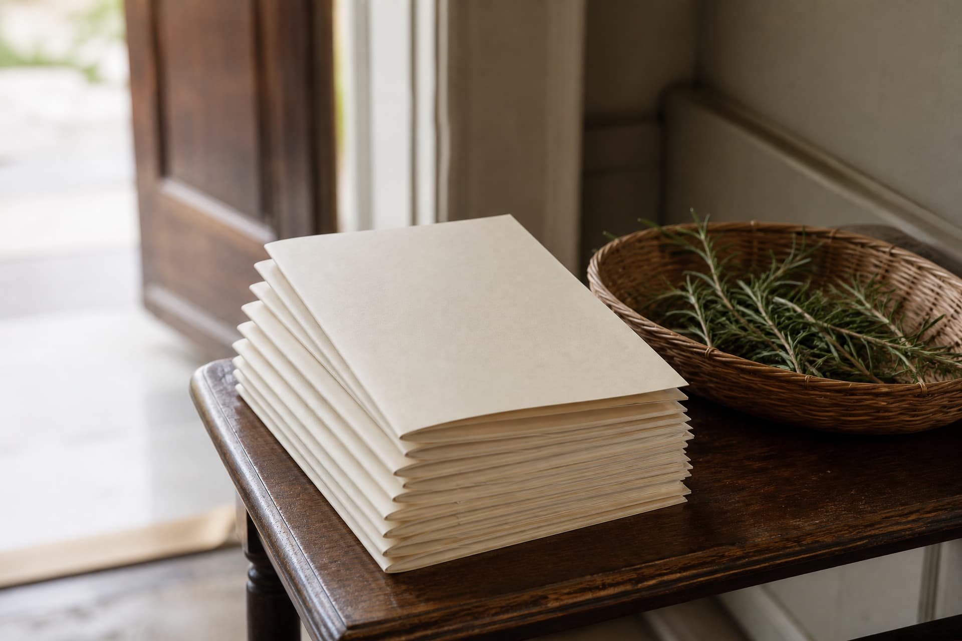 A neat stack of folded cream memorial programs on a small side table just inside a doorway, the top one slightly askew, with a shallow basket of loose rosemary sprigs beside them