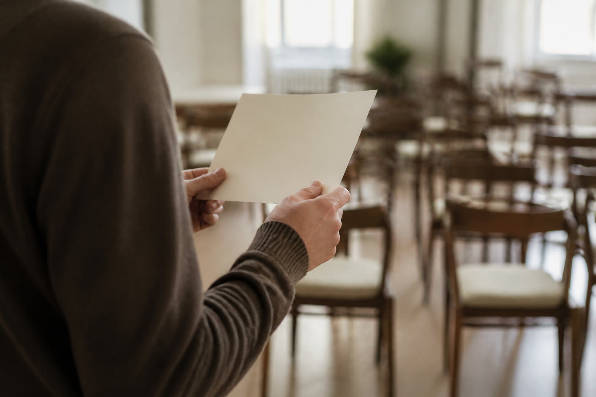 A single sheet of cream card held in two hands at chest height, seen from behind and to the side with the reader's head out of frame, rows of empty wooden chairs softly out of focus beyond