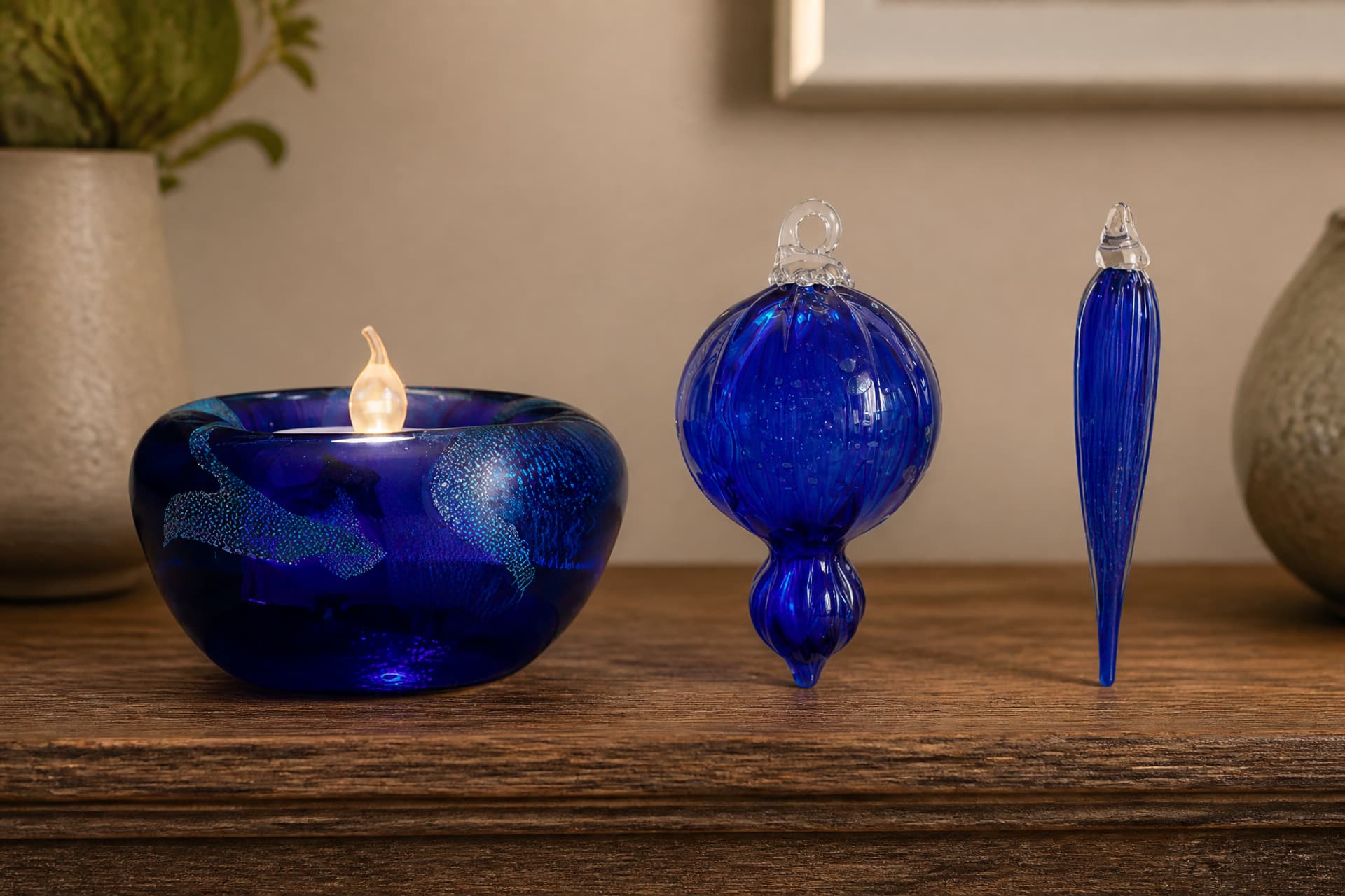 Three pieces of deep blue glass on a wooden sideboard, a lit candle bowl with iridescent swirls, a round hanging bauble and a slender tapered icicle