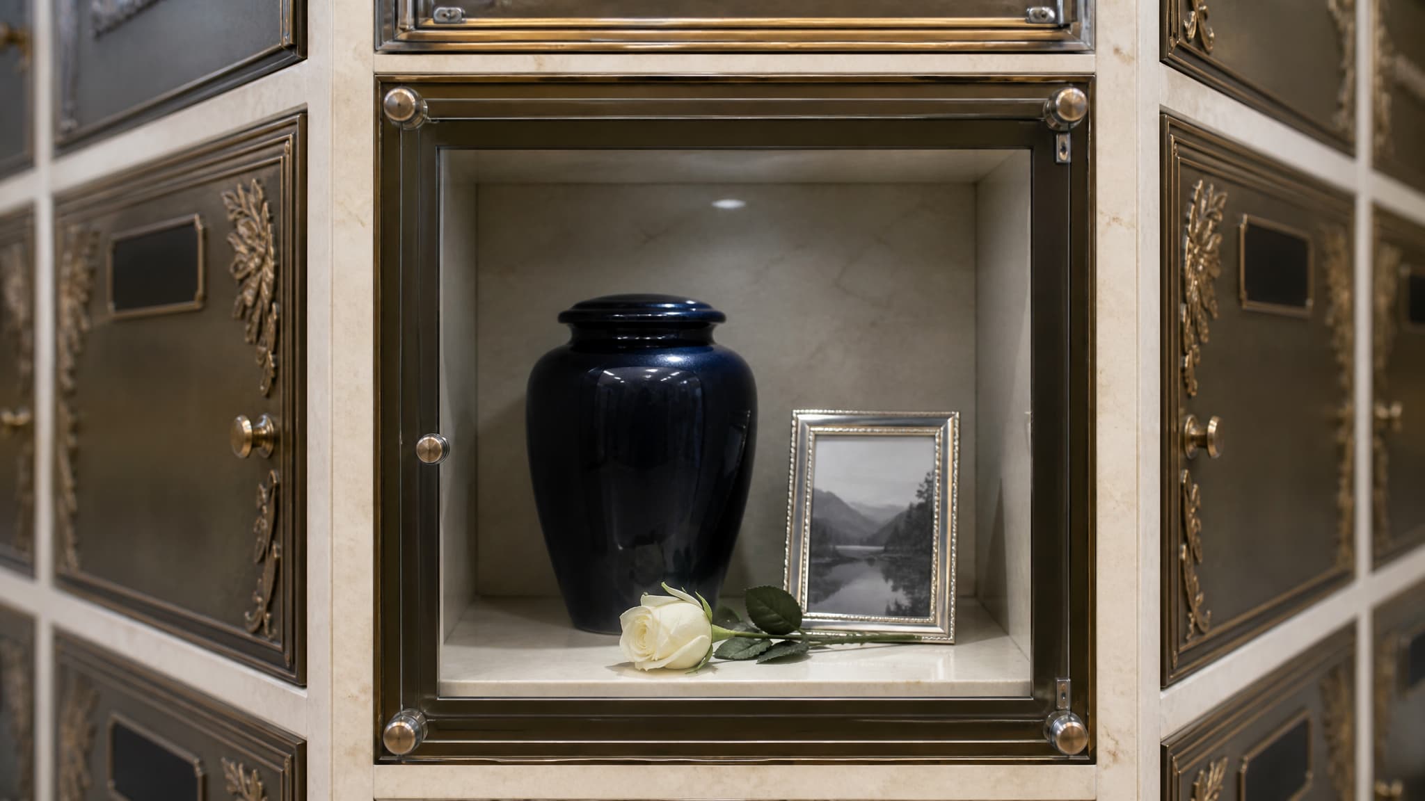 A glass-fronted columbarium niche photographed straight on, holding a dark ceramic urn with a small framed photograph beside it and a single artificial white rose, with rows of engraved bronze niche fronts blurred to either side