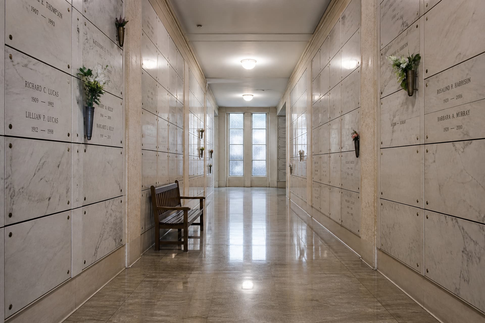 The interior of an indoor mausoleum corridor lined floor to ceiling with marble niche fronts, a polished stone floor reflecting soft ceiling light and a simple wooden bench against one wall