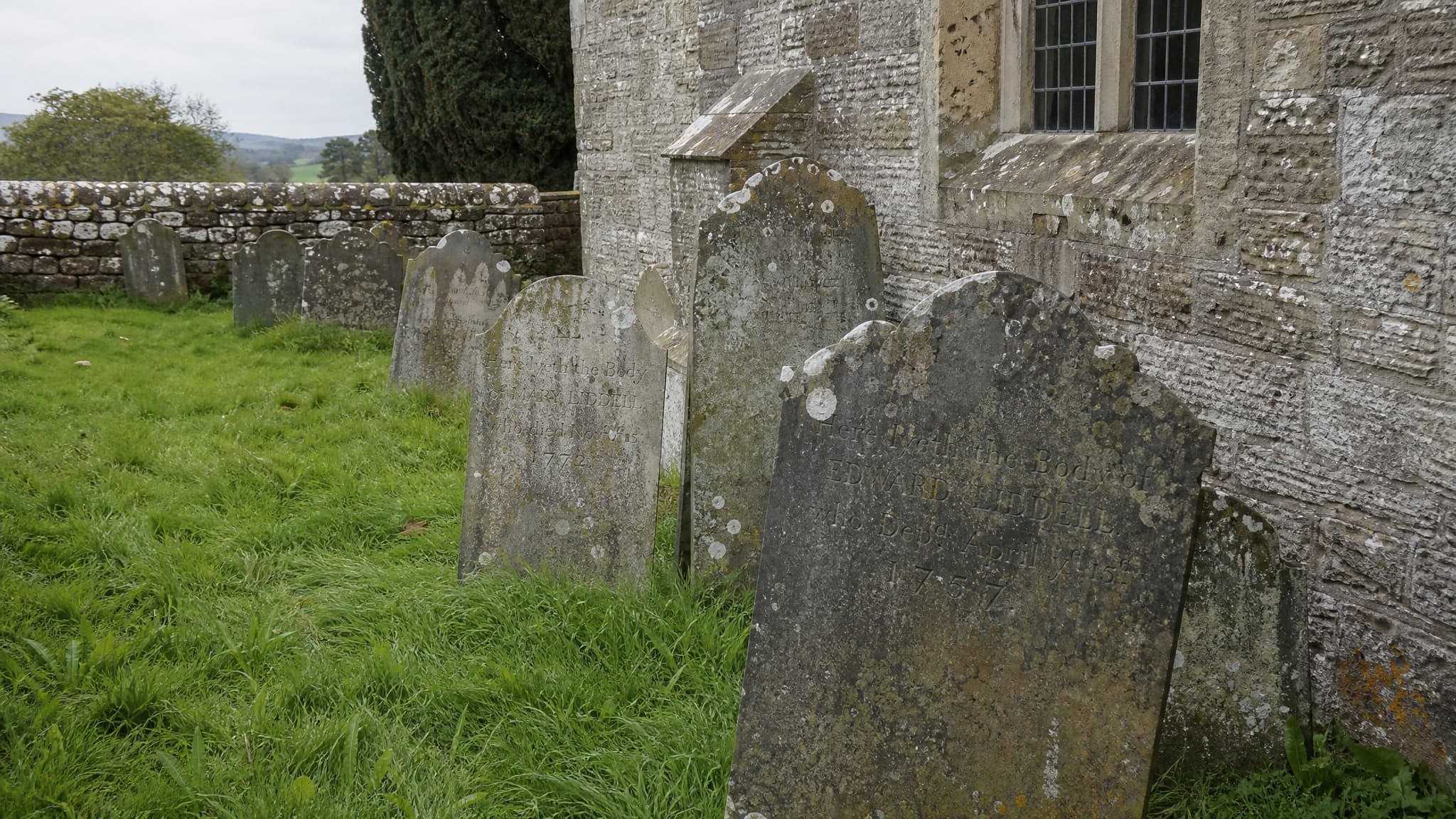 A split view of two burial grounds photographed on the same overcast morning: on the left a small crowded churchyard of leaning slate stones pressed against a stone church wall, on the right a wide open lawn cemetery with widely spaced granite monuments and mown avenues