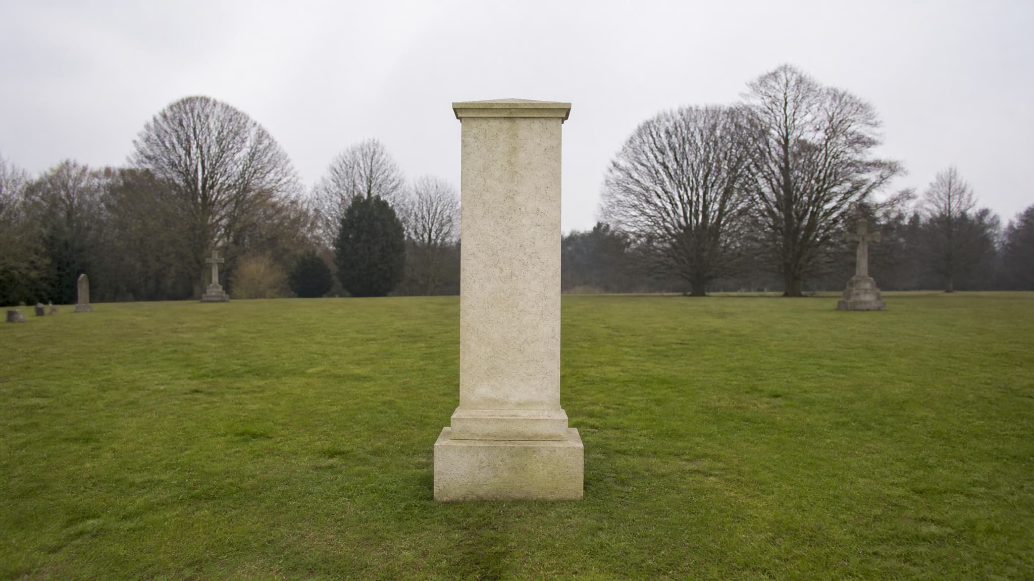 A plain pale stone memorial pillar standing alone on mown grass under an overcast sky, no grave plot beneath it and bare trees at a distance behind