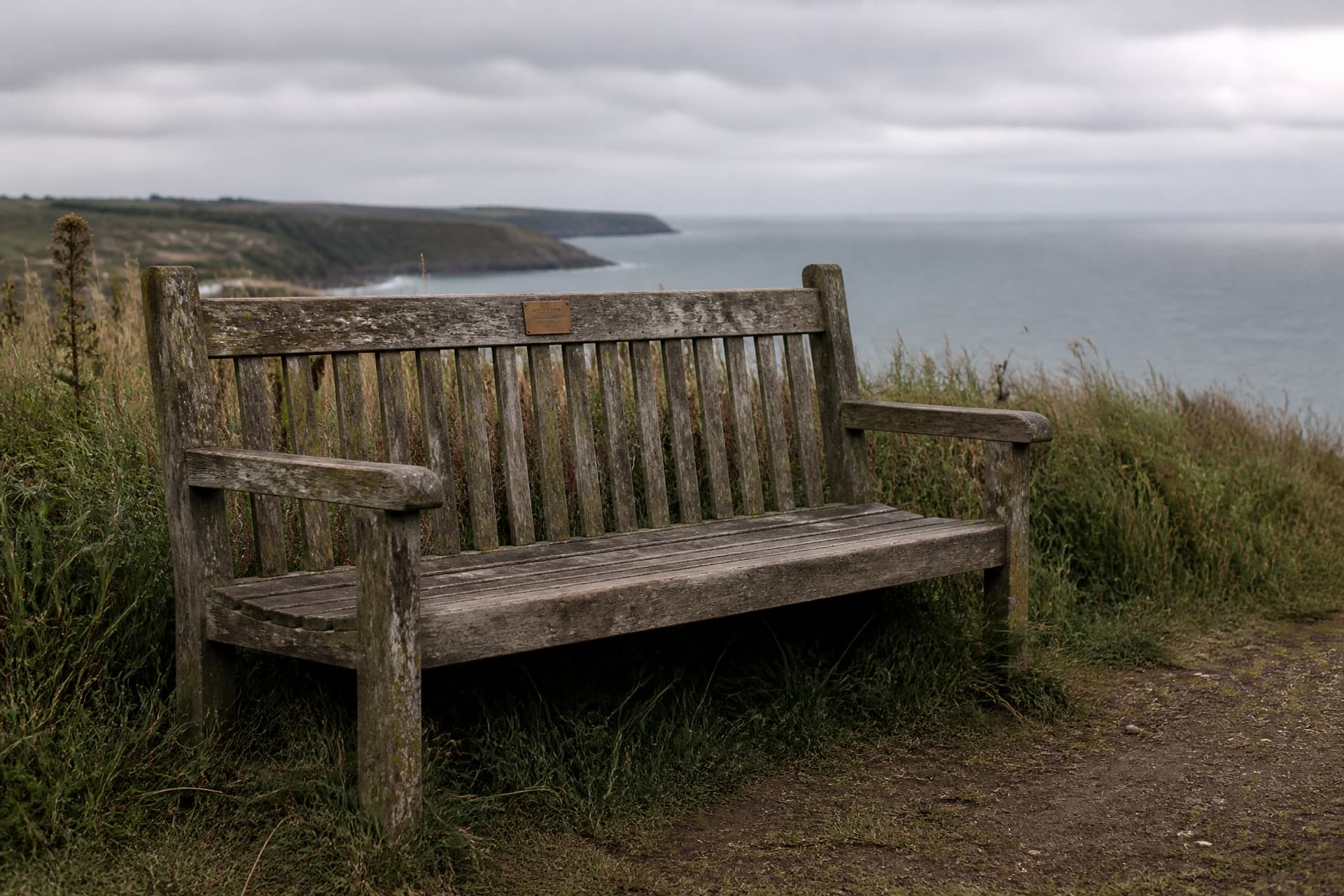 A weathered wooden memorial bench on a grassy coastal path with a small brass plaque fixed to its backrest, sea and low cloud beyond the cliff edge
