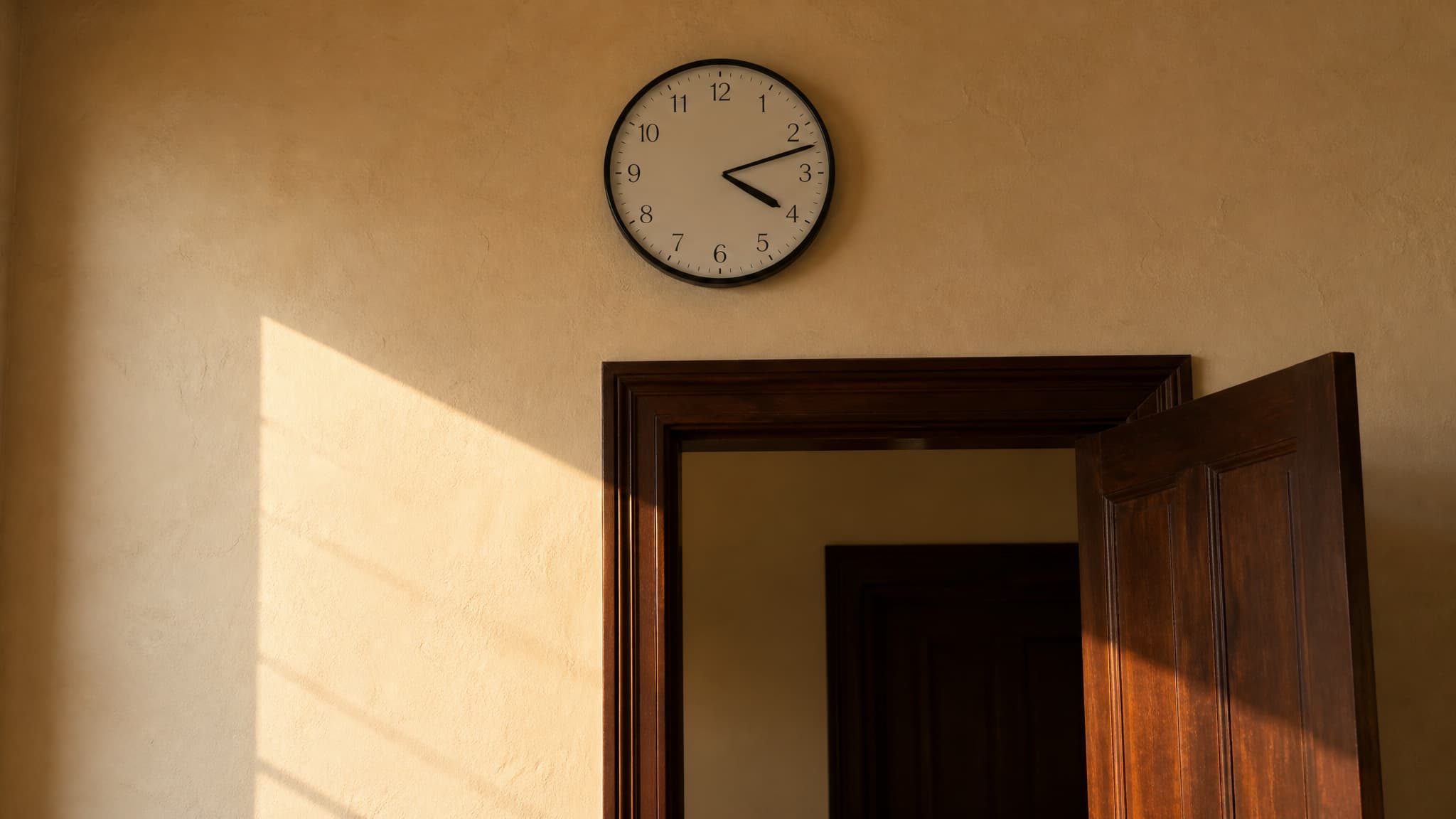 A plain round wall clock with black numerals and no lettering, reading ten past four, high on a warm cream plaster wall above a dark panelled doorway standing open, late afternoon sun slanting across the wall from the left