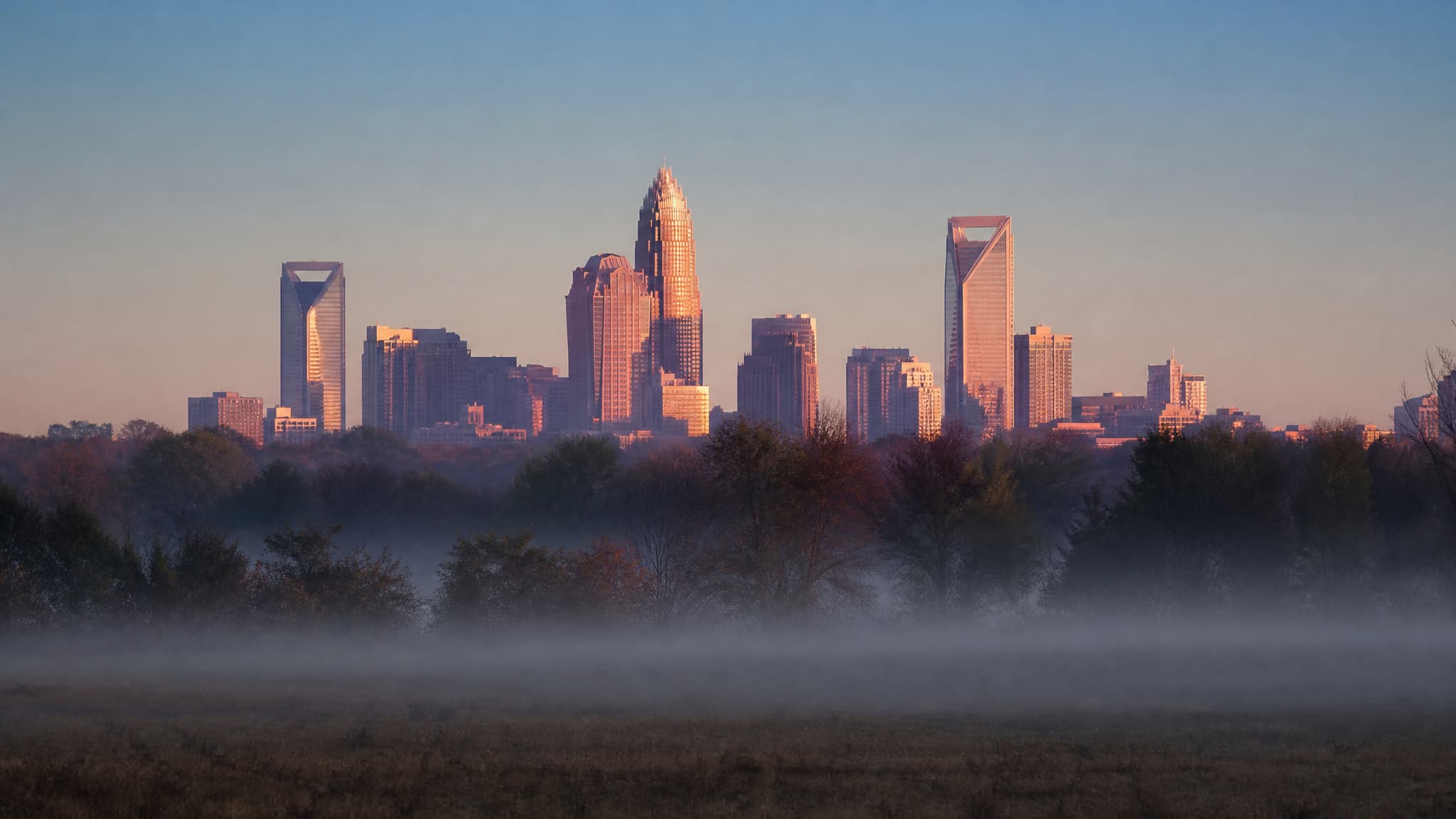The Charlotte skyline seen from open farmland several miles away at dawn, a compact cluster of glass and stone towers lit pink and gold on the horizon with mist lying across the fields in the foreground