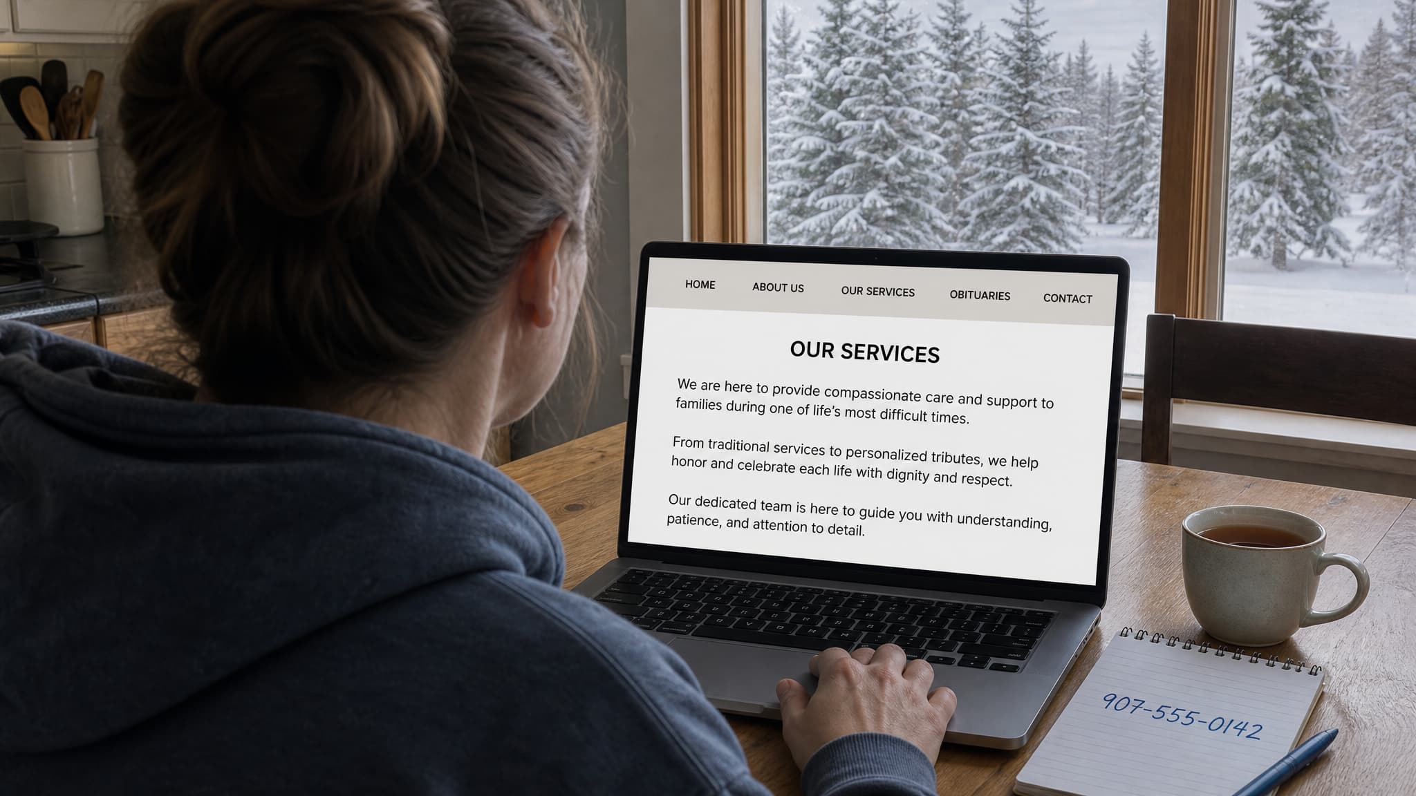A kitchen table in an Alaskan home with a laptop open to a funeral home website showing no prices, a notepad with a phone number written on it, and snow-covered spruce visible through the window
