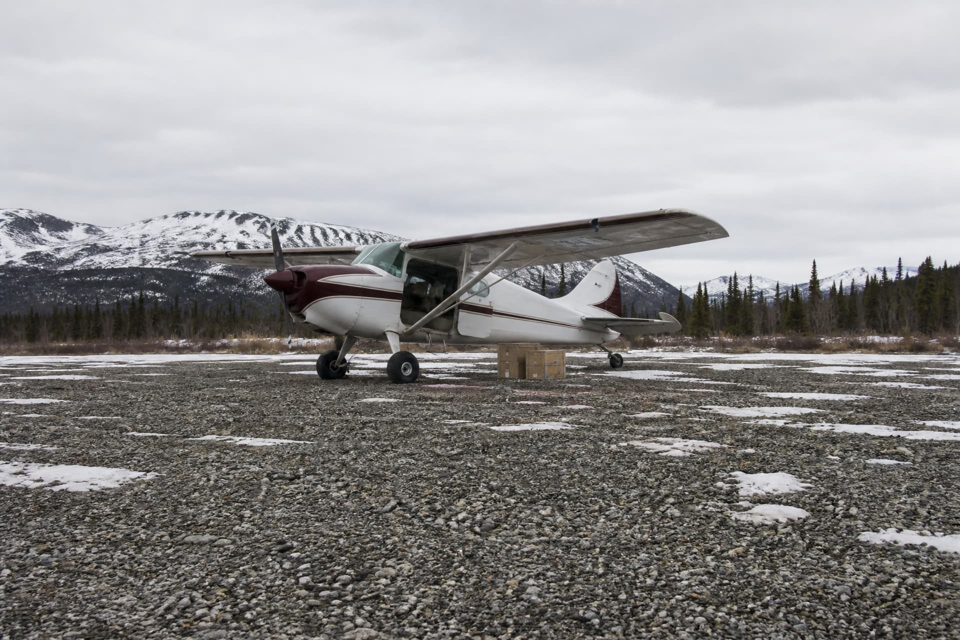 A small aircraft on a gravel airstrip in rural Alaska with mountains behind it, cargo being loaded in flat winter light
