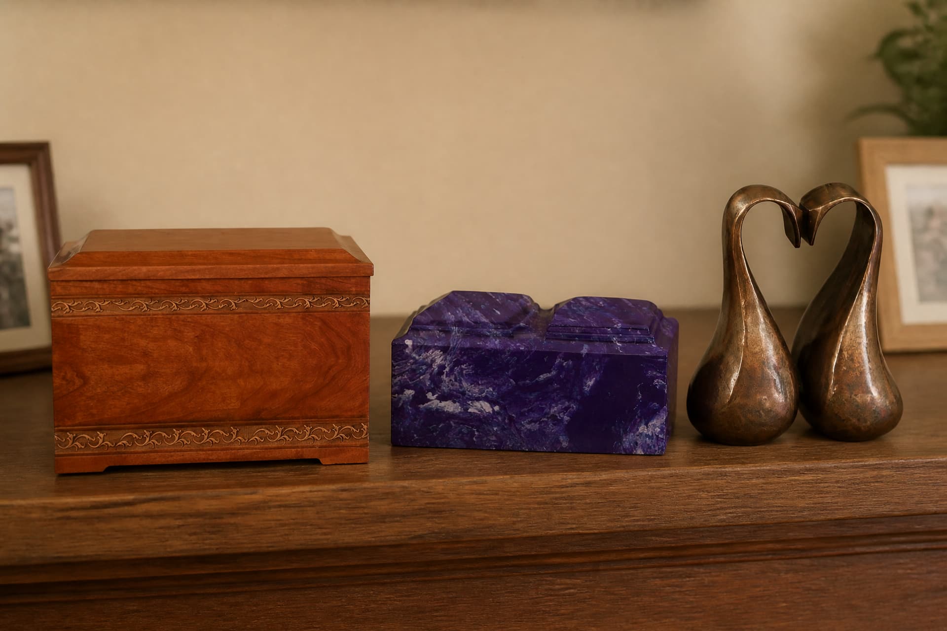 Three companion cremation urns in a row on a wooden mantel โ a cherry wood chest with carved scroll bands, a wide purple marbled stone box with two lids, and a pair of bronze swans meeting in a heart