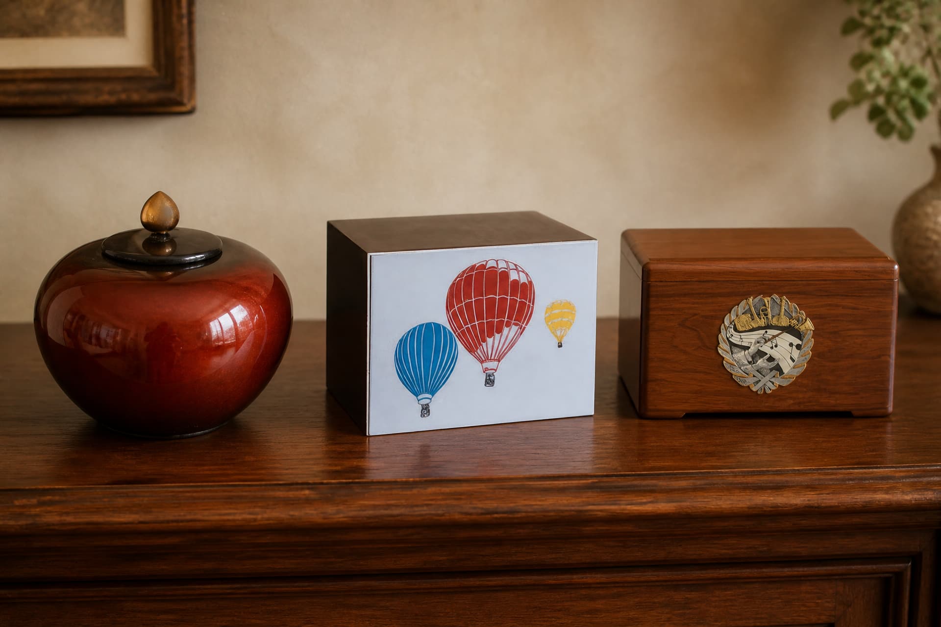Three distinctive cremation urns on a wooden sideboard โ a deep red glazed jar with a gold finial, a dark wood box painted with three hot air balloons, and a walnut box with a musical medallion